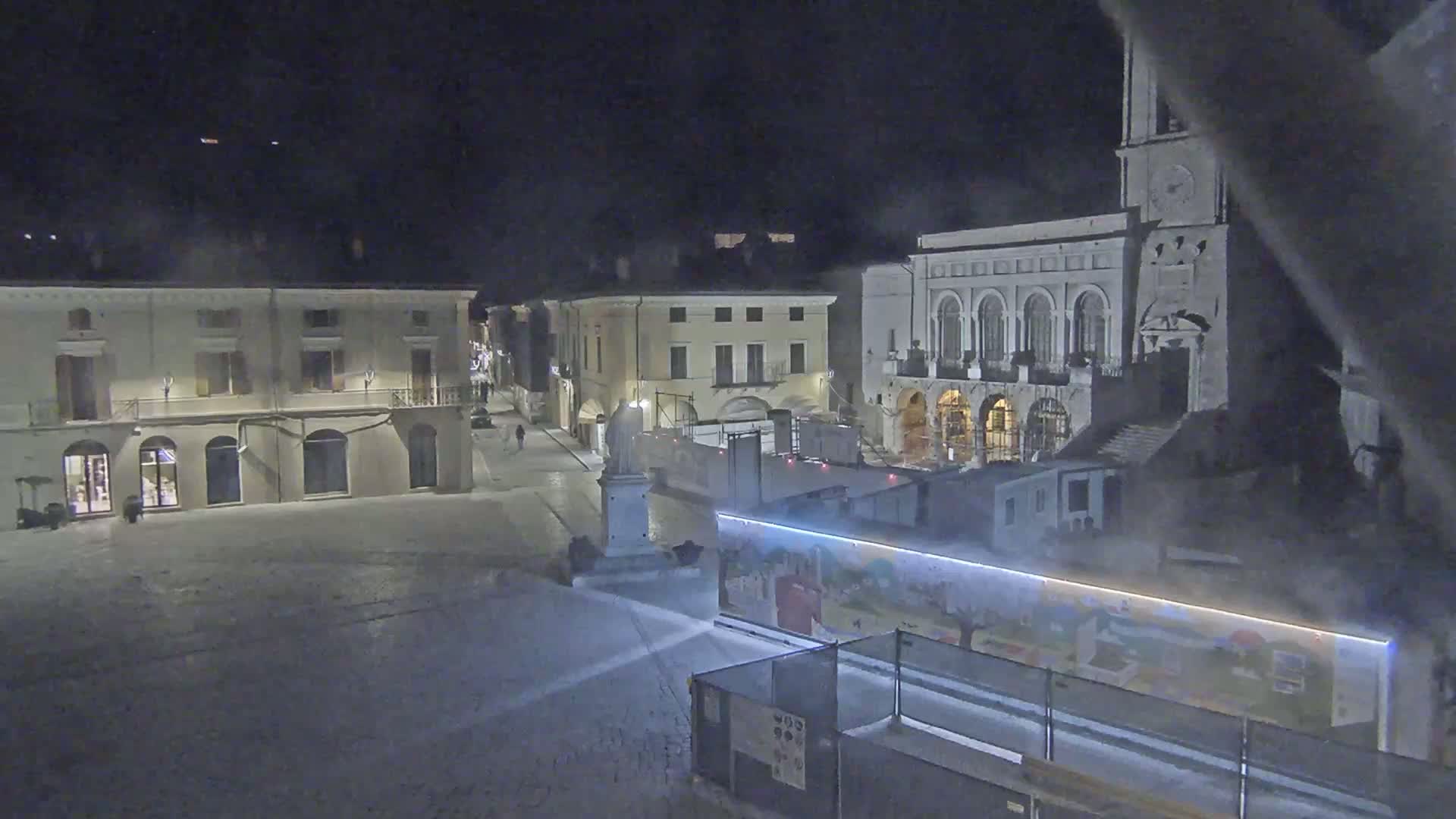 A mostly empty town square at night, under a dark sky, is partially obscured by construction barriers and illuminated by artificial lights.