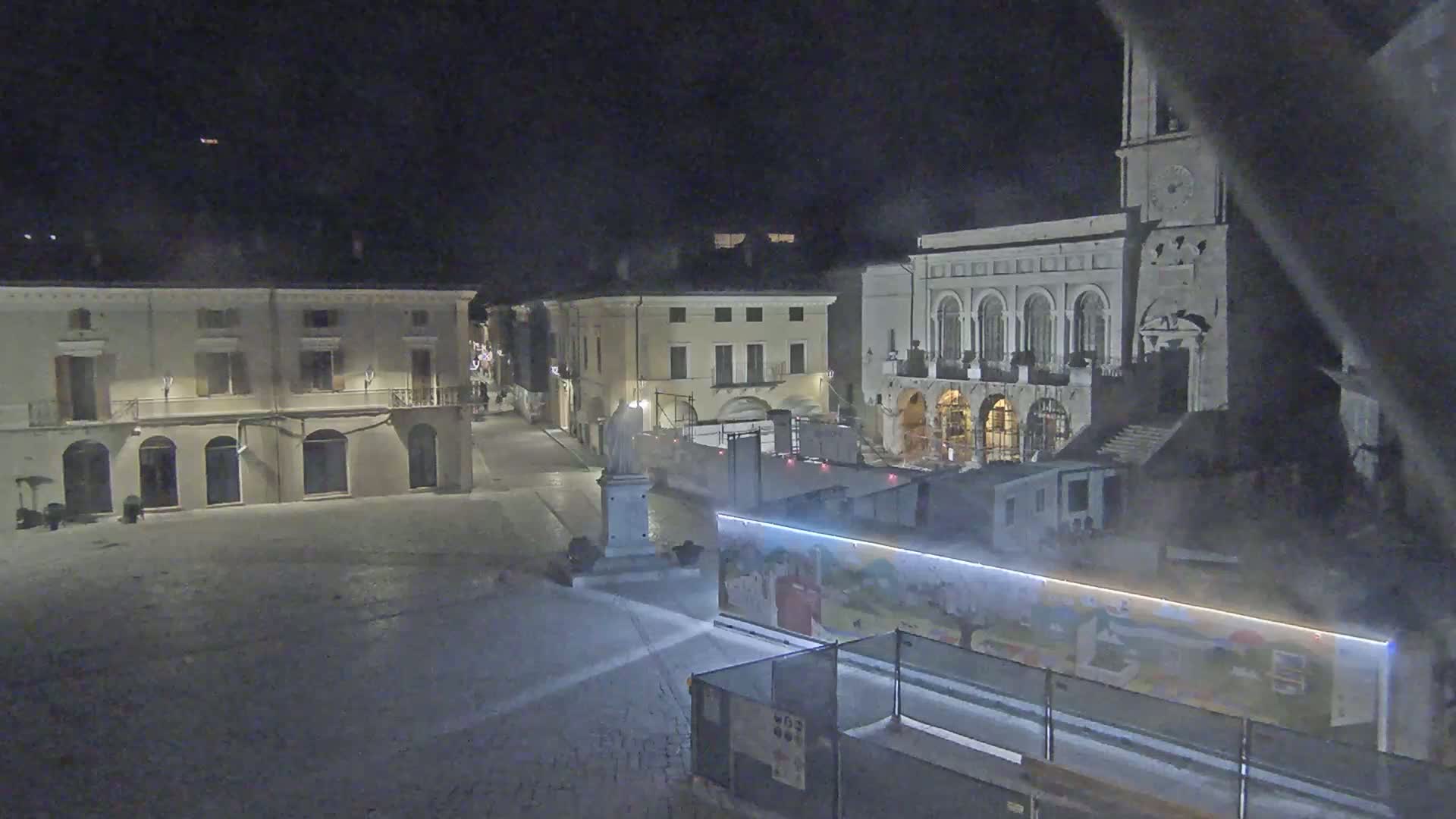 A nighttime view of a town square, with several buildings, a statue, and construction barriers under dark skies.