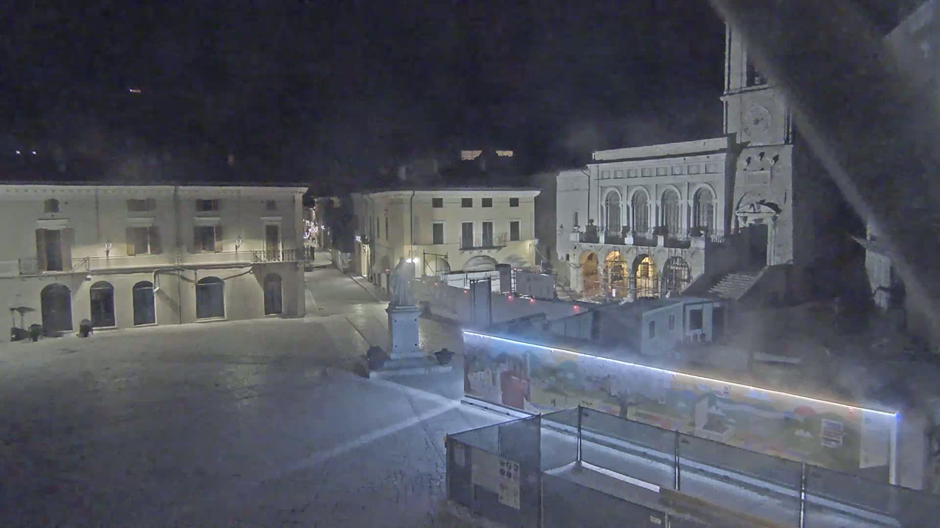 A snow-covered town square at night is surrounded by buildings, one of which is a large church with a clock tower.