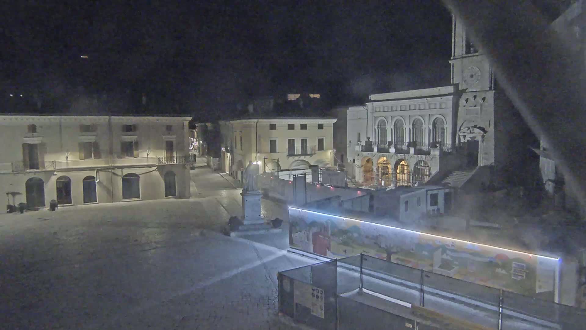 A nighttime view of a town square with several buildings, including a large church, partially obscured by construction barriers and mist.