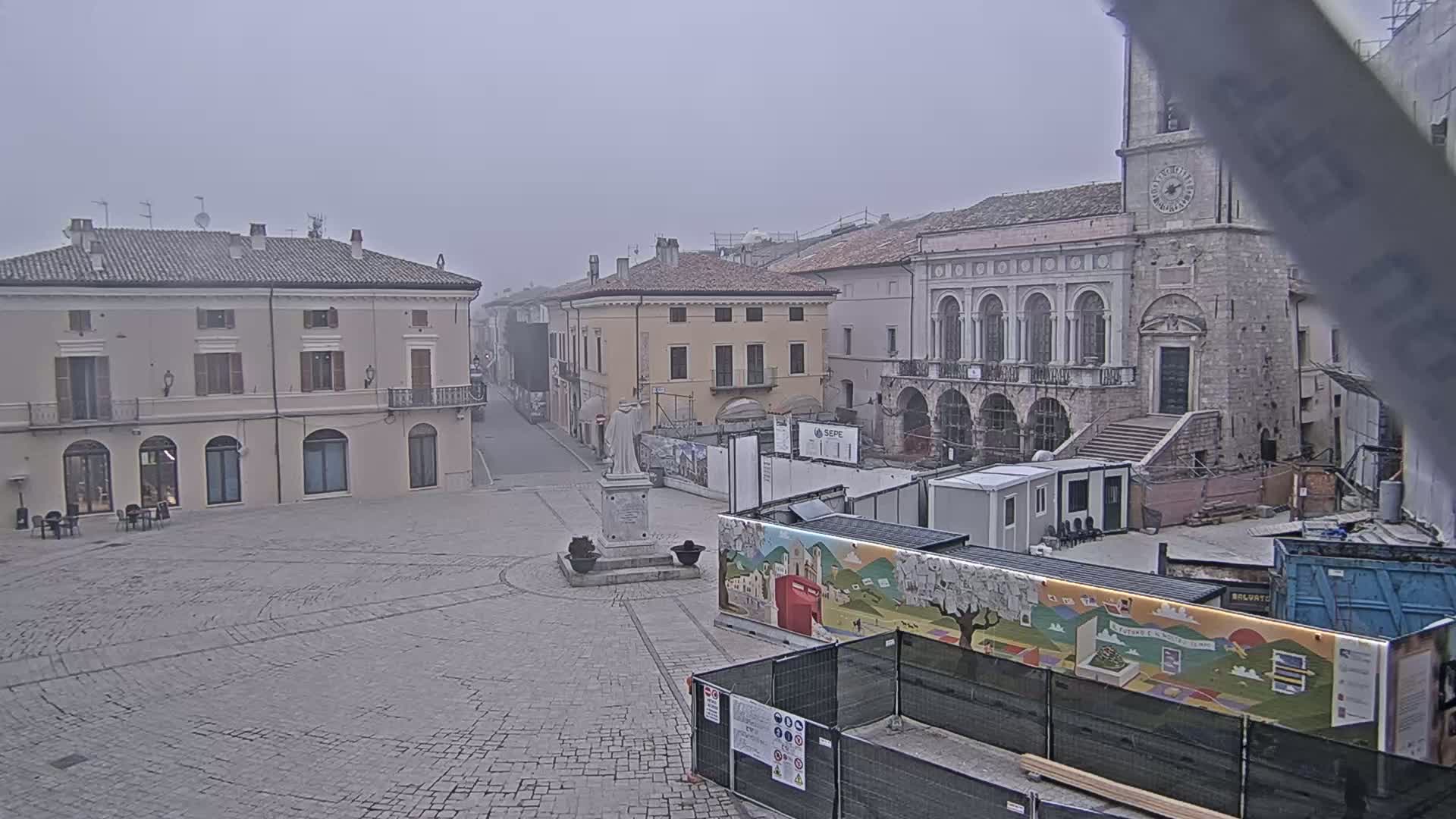 A mostly empty cobblestone town square is shown under an overcast sky, with several buildings surrounding it, one of which is under construction.