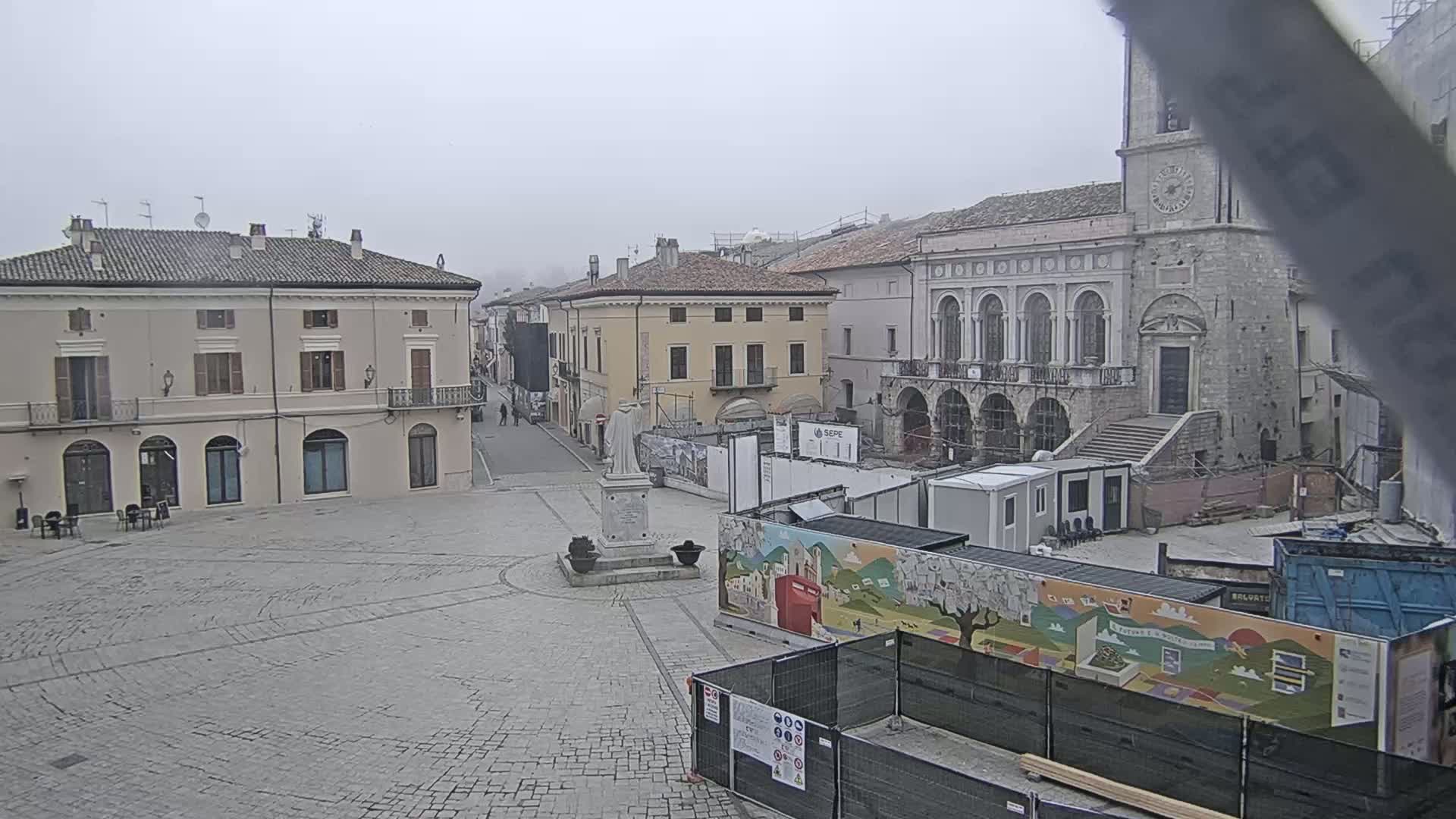 A town square is shown on an overcast day, with construction underway near a large stone building and a statue in the center of the square.
