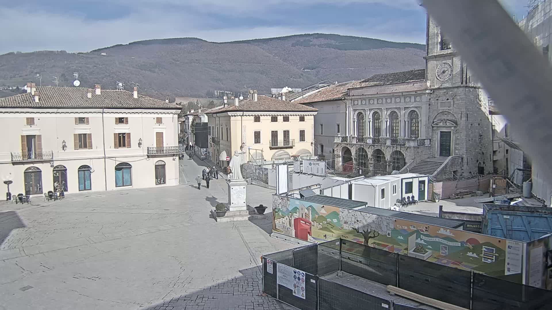 A town square with several buildings, a statue, and construction underway, under a clear, sunny sky with mountains in the background.