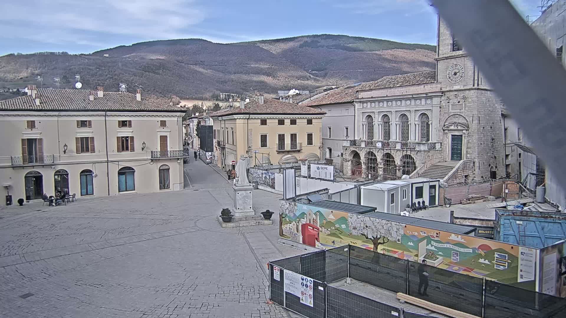 A mostly empty town square with several buildings, including a large stone building with a clock tower, is under partly cloudy skies; construction is underway in part of the square.