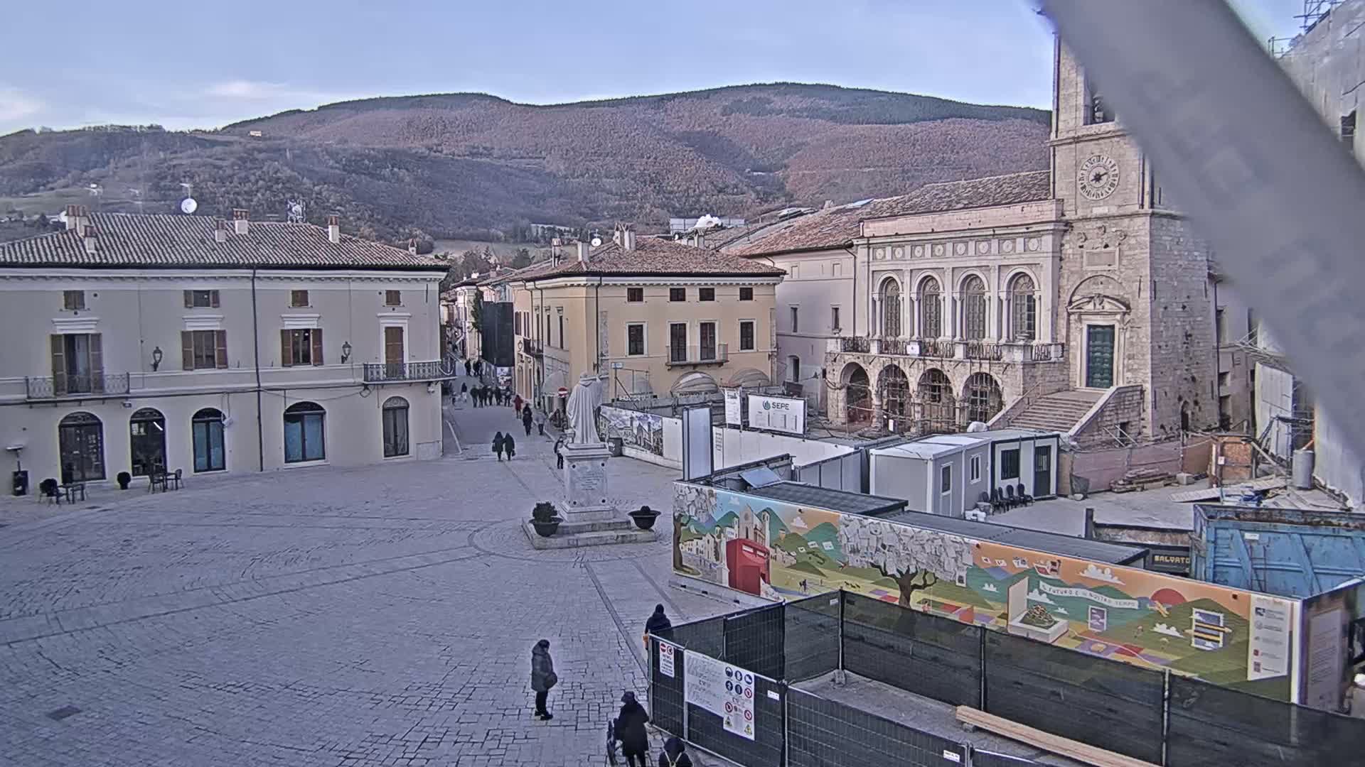 A town square with several buildings, a statue, and construction underway, is viewed on a clear, cool day against a backdrop of distant mountains.