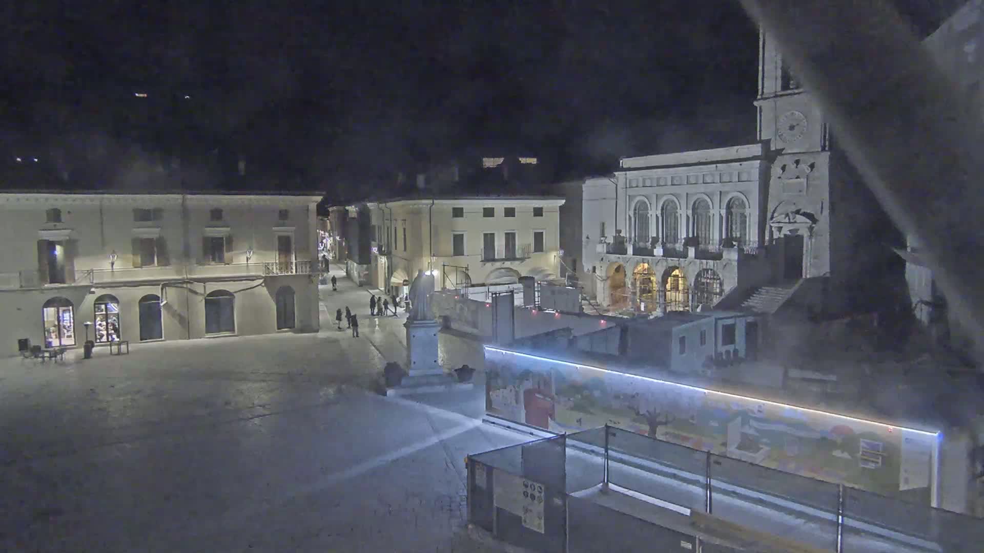 A snow-covered town square at night, with several buildings, a statue, and construction barriers illuminated by artificial light.