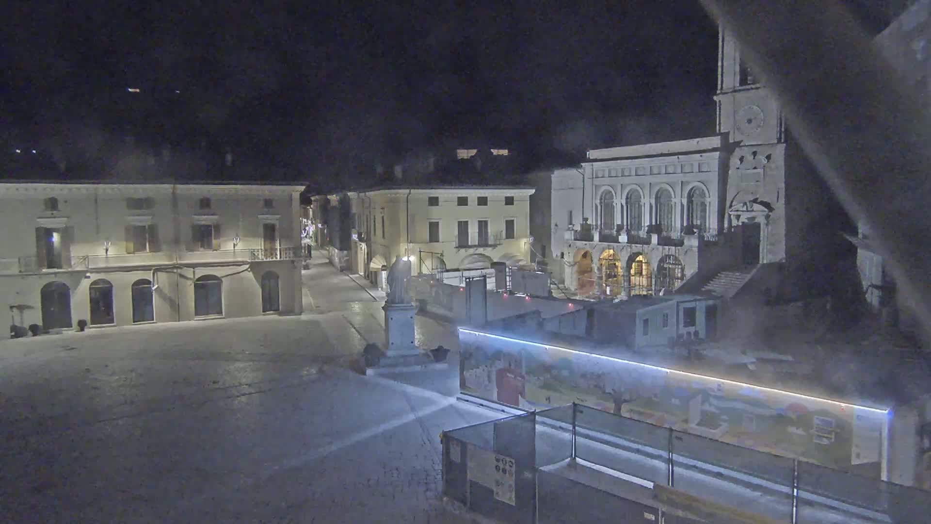 A nighttime view of a snow-covered town square with several buildings, a statue, and construction barriers.
