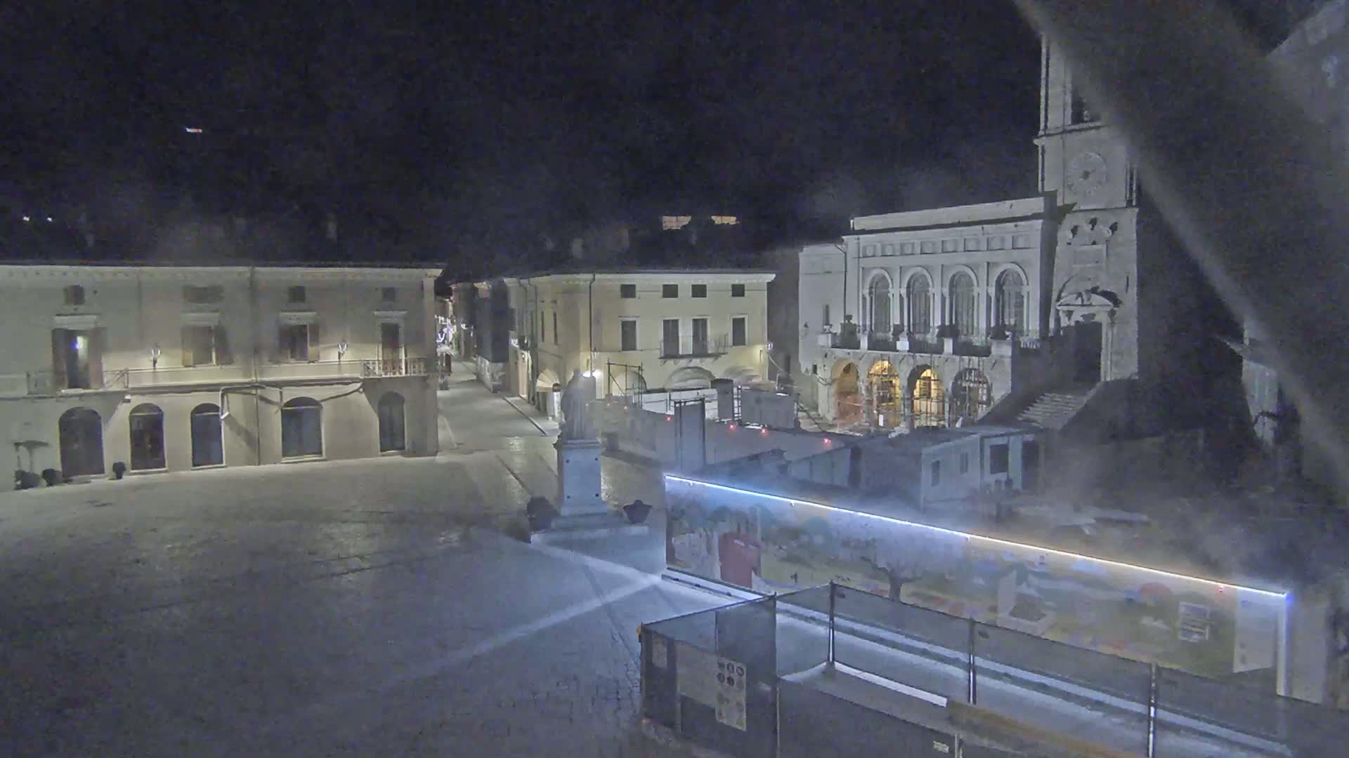 A snow-covered town square at night is partially obscured by construction barriers and fog, with classical-style buildings visible in the background.