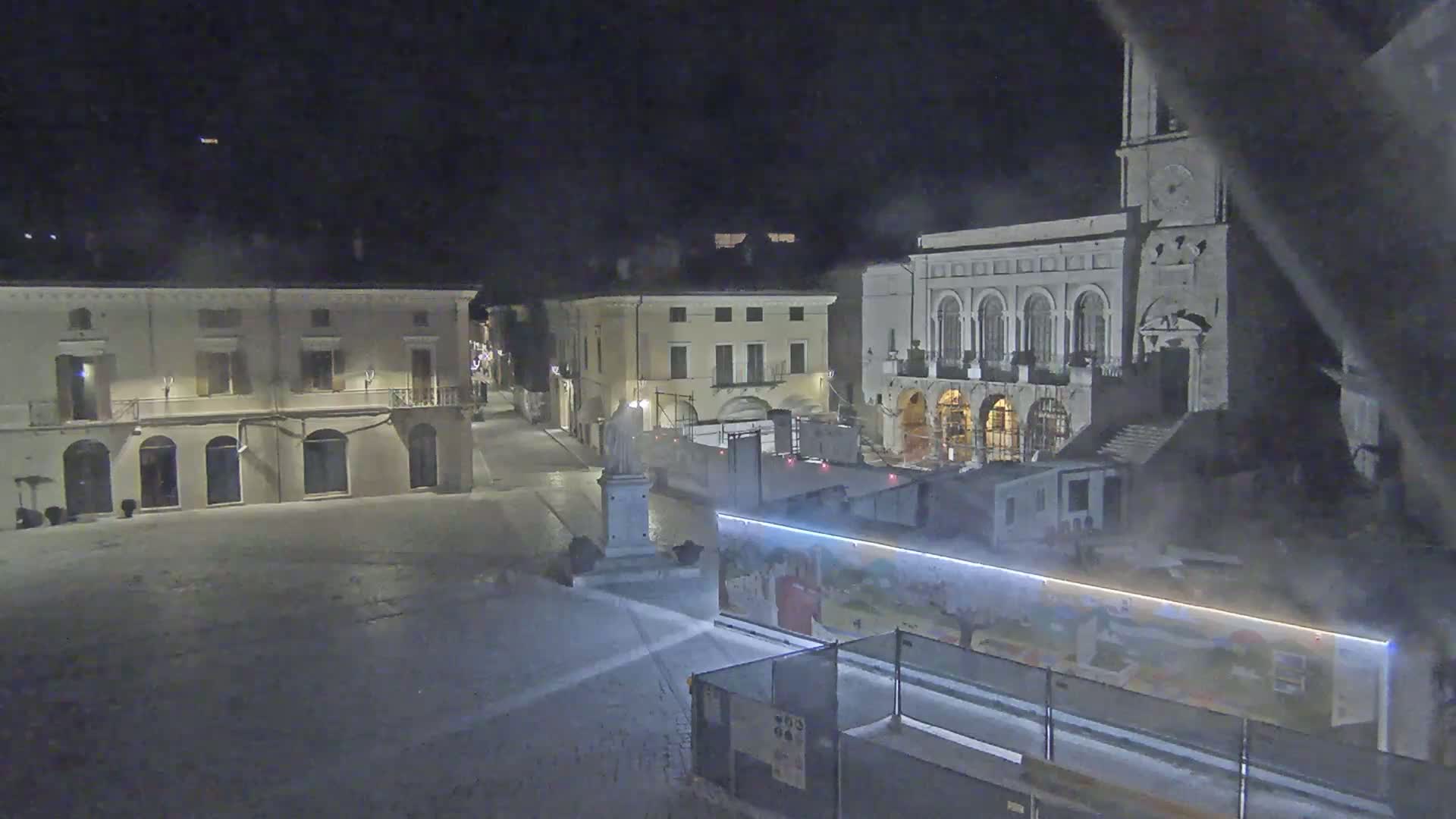 A snow-covered town square at night is seen, with several buildings illuminated, including a large building with arched windows and a clock tower.