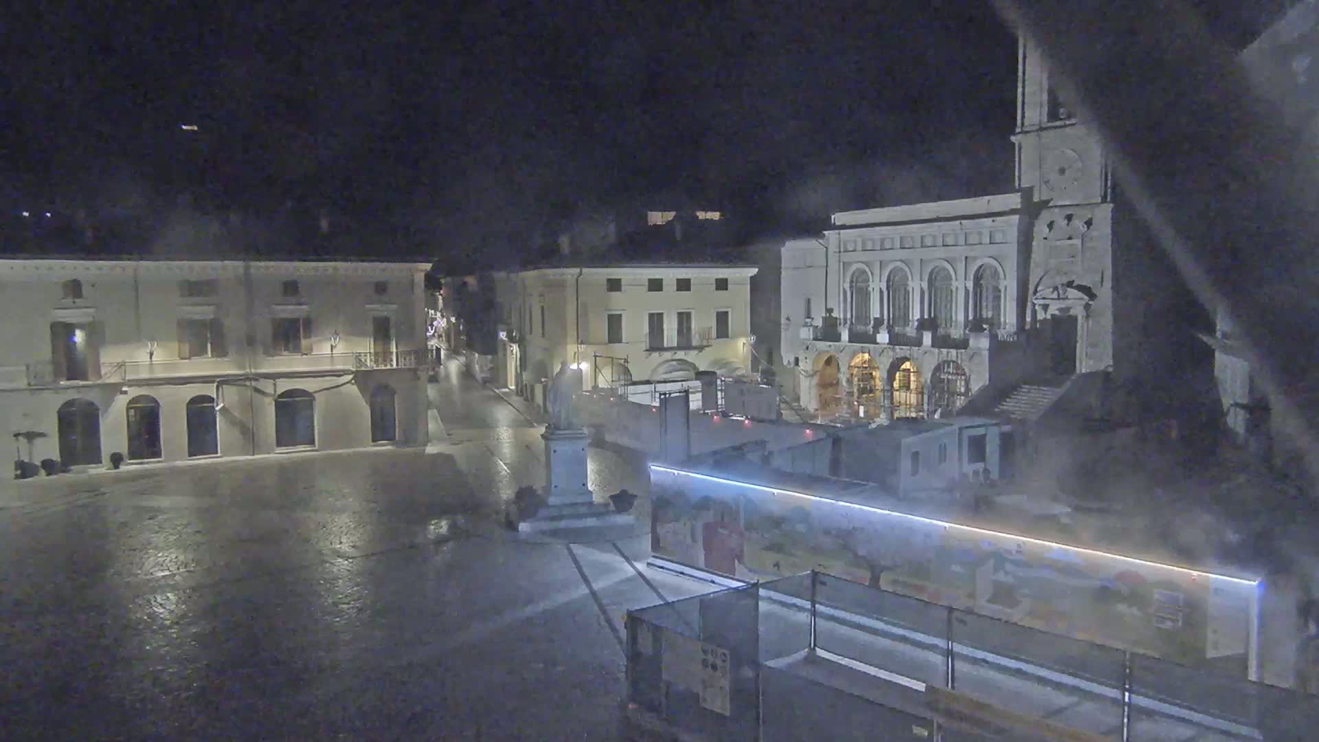 A nighttime view of a town square, wet from rain, showing several buildings, a statue, and construction barriers.