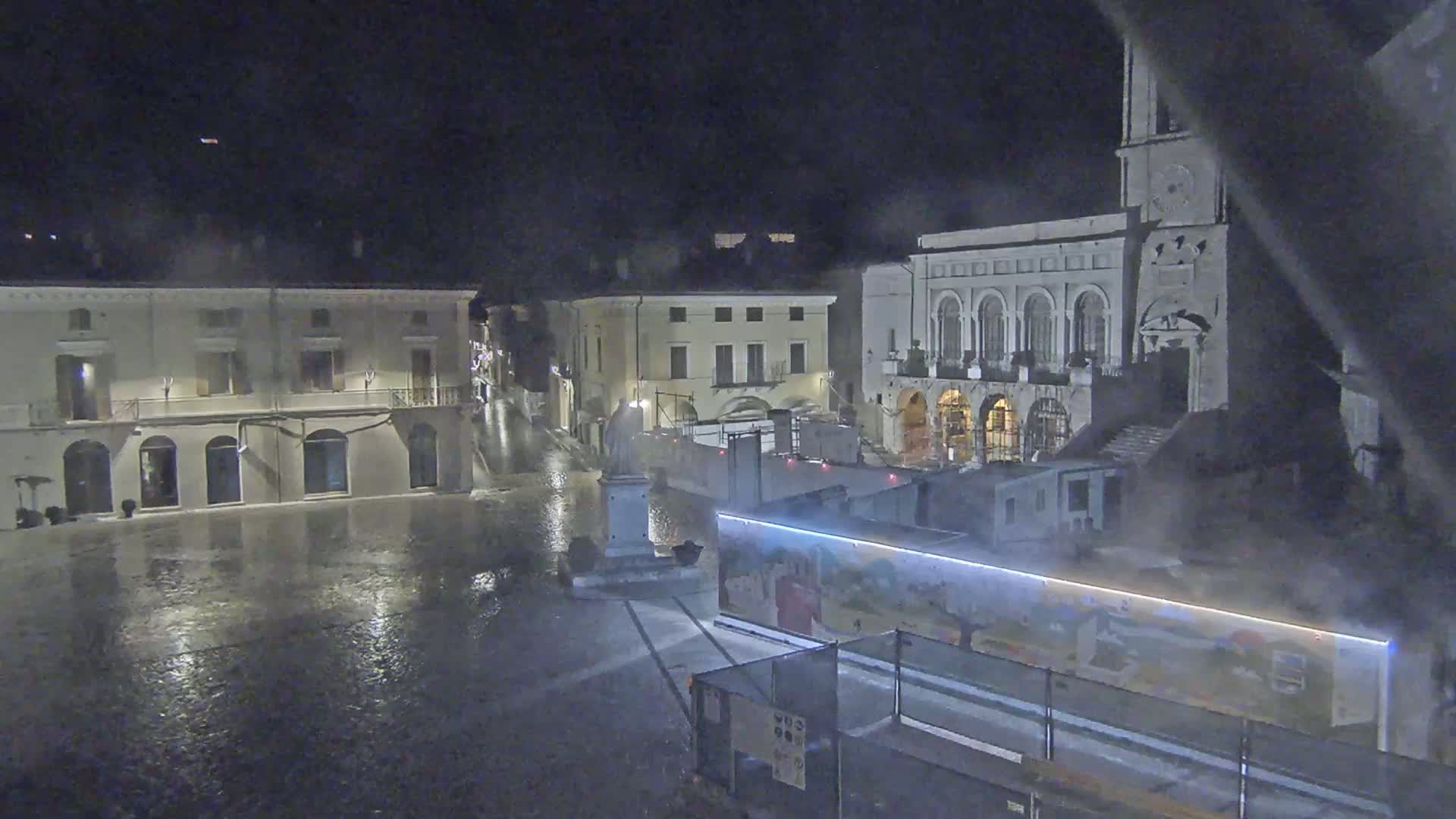 A rain-soaked town square at night is surrounded by buildings, one of which has a clock tower, with construction barriers and equipment present in the square.