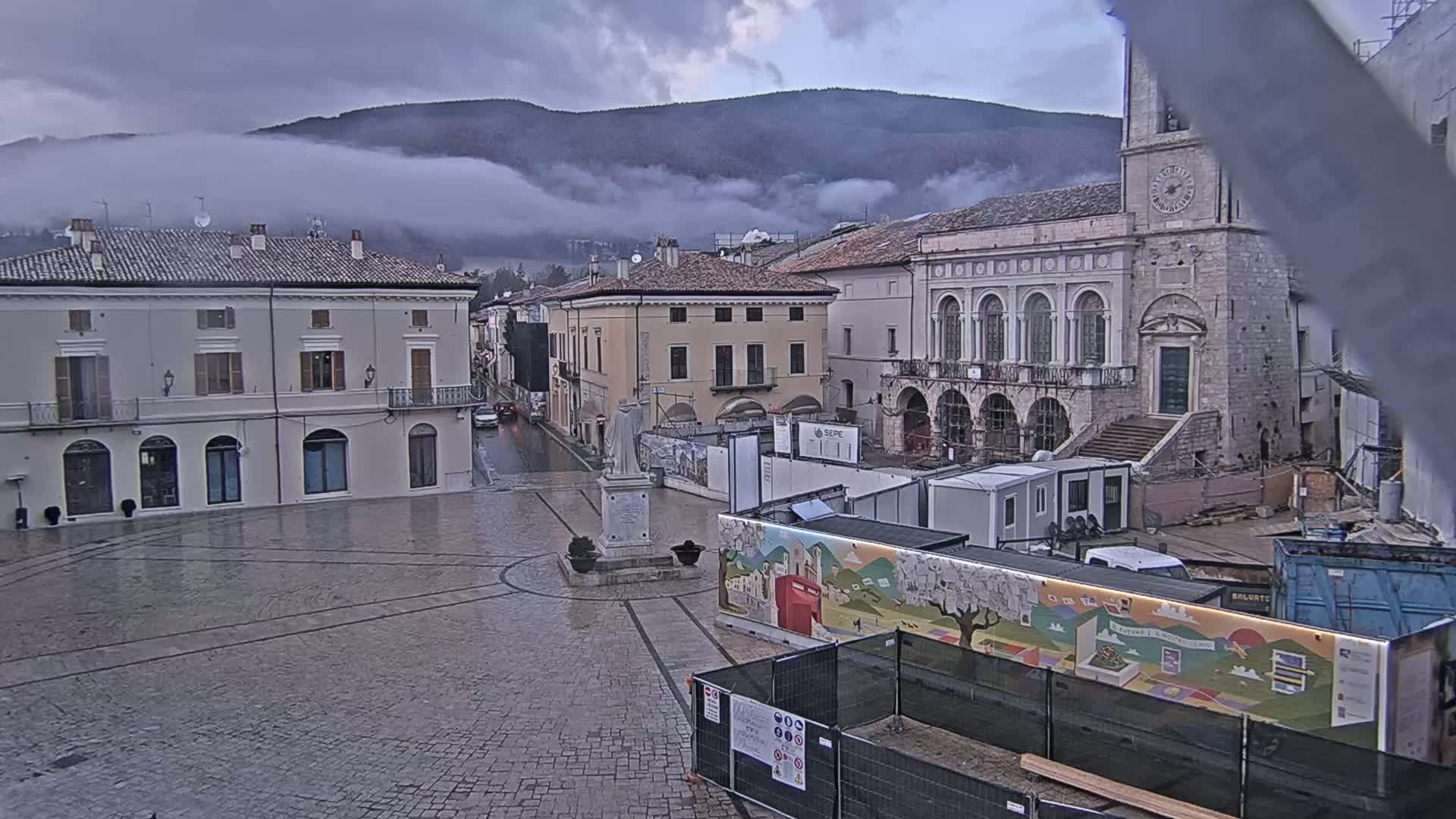A town square with several buildings, a statue, and construction barriers, under a cloudy sky with fog in the mountains in the background.