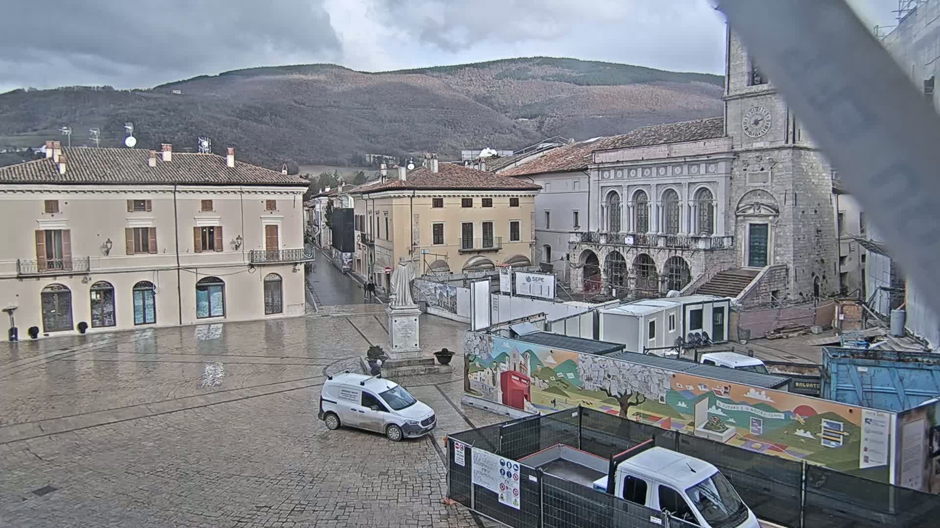 A town square with several buildings, a statue, and construction equipment under an overcast sky.