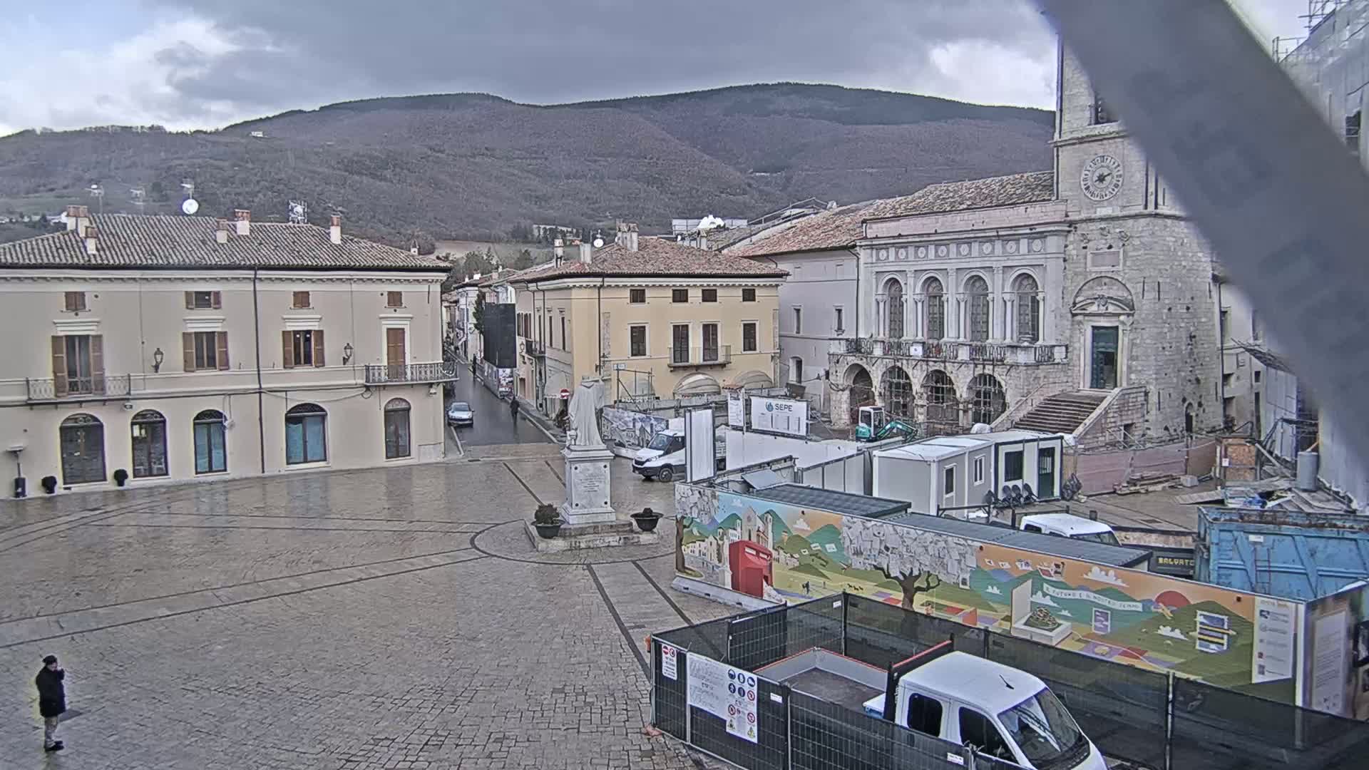 A wet, cobblestone town square is under a cloudy sky, with several buildings surrounding it, one of which is undergoing construction, and mountains in the background.