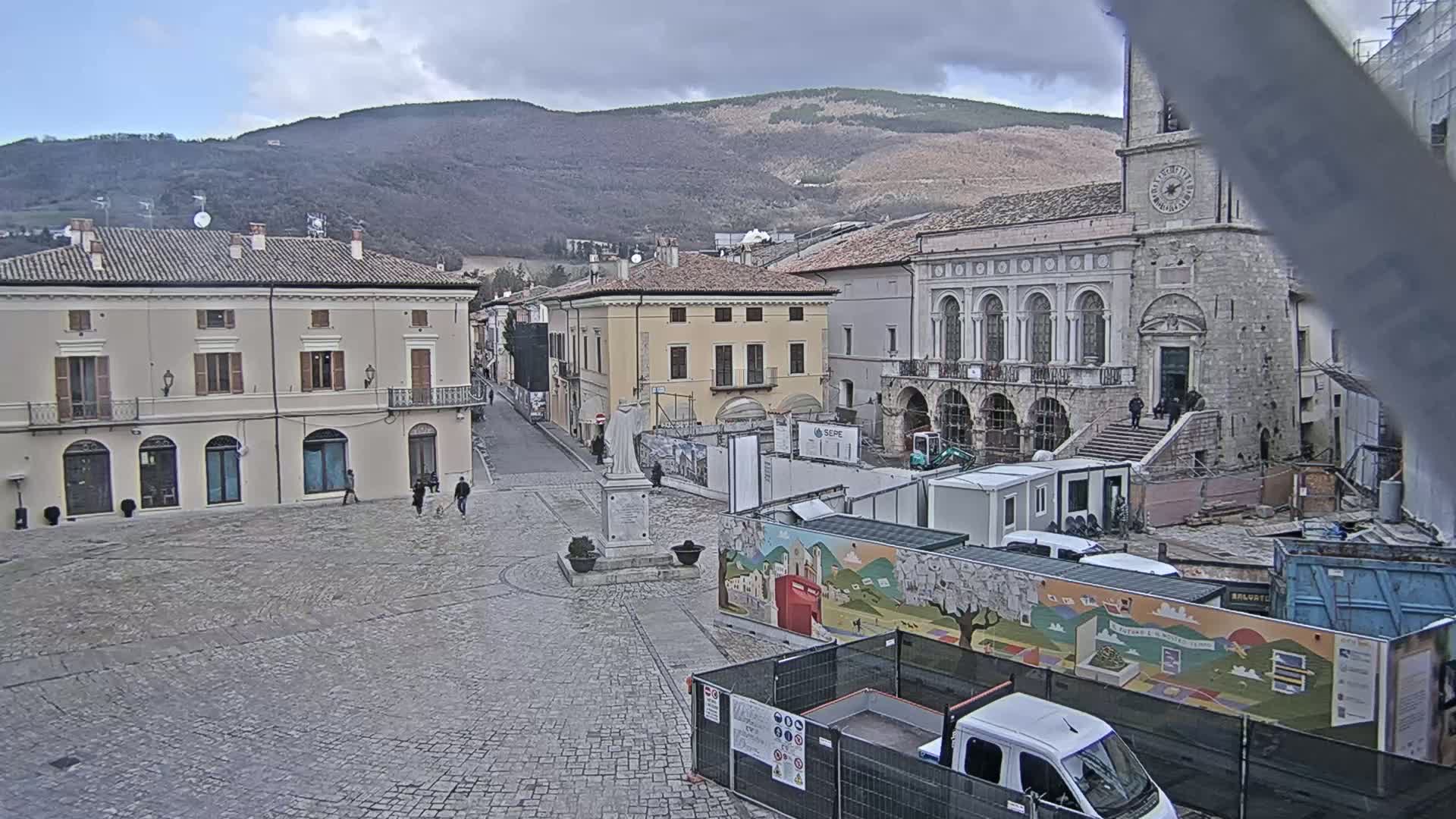 A town square under a cloudy sky features several buildings, a statue, construction equipment, and a few pedestrians.