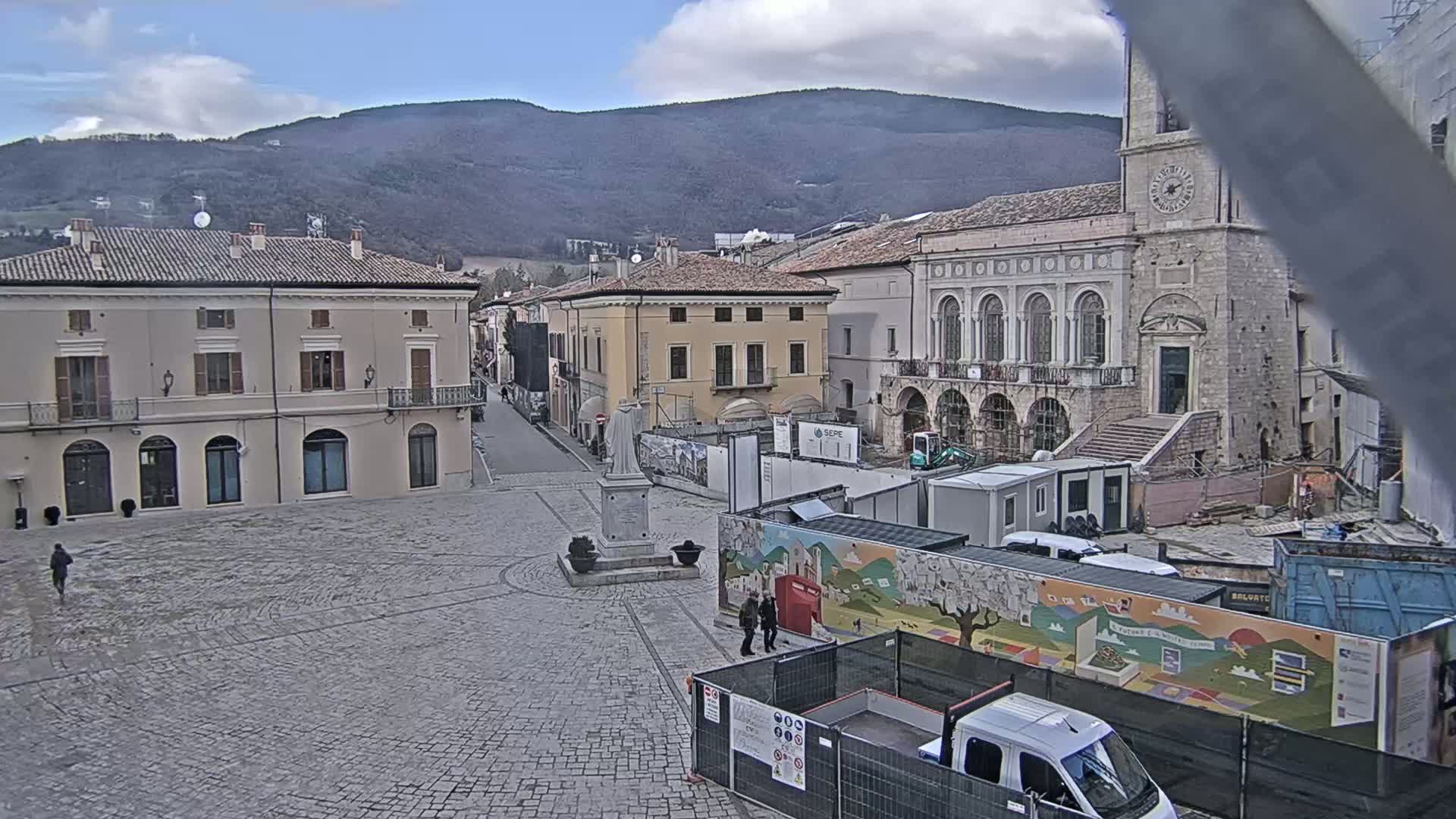 A cobblestone town square is under construction, with several buildings surrounding it, including a large stone building with arched windows, and mountains in the background under a partly cloudy sky.