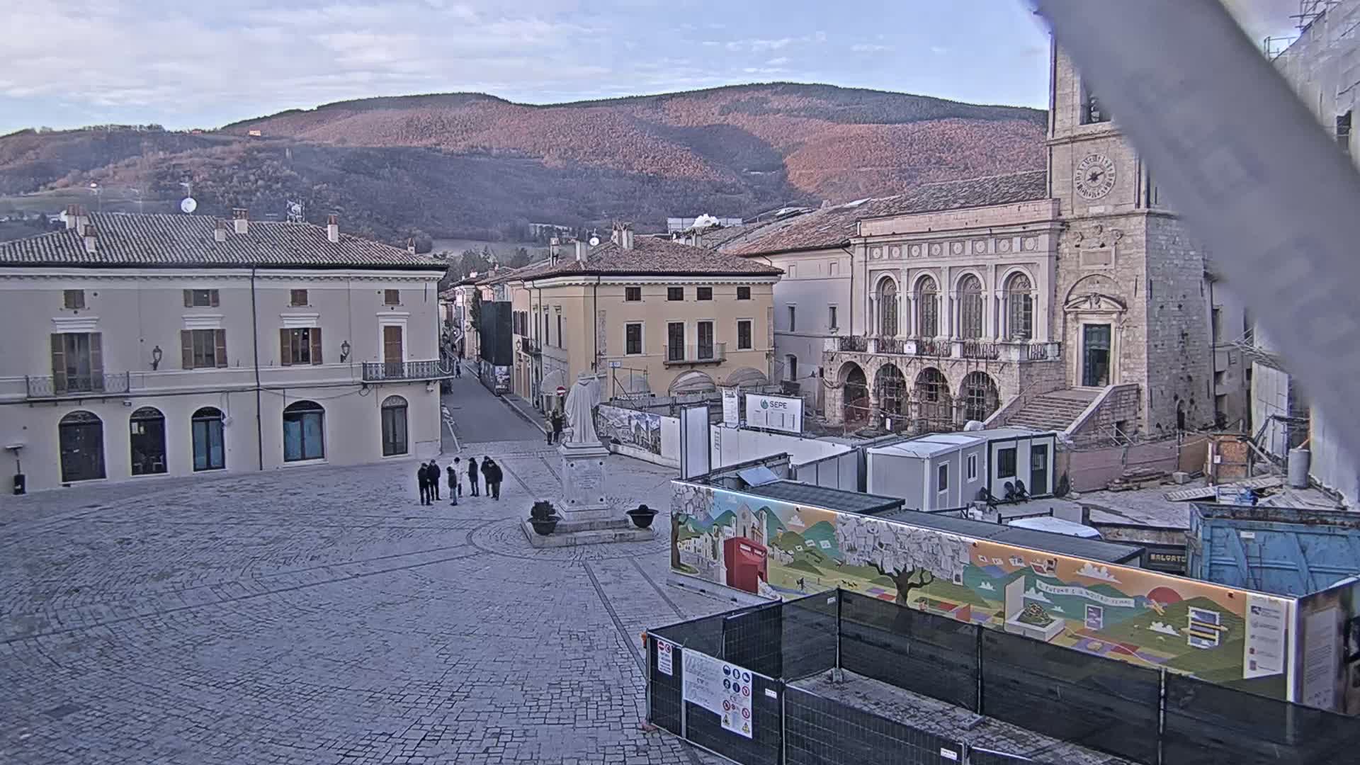 A town square under a partly cloudy sky, with several buildings surrounding it, including a large stone church, and construction underway in the center.