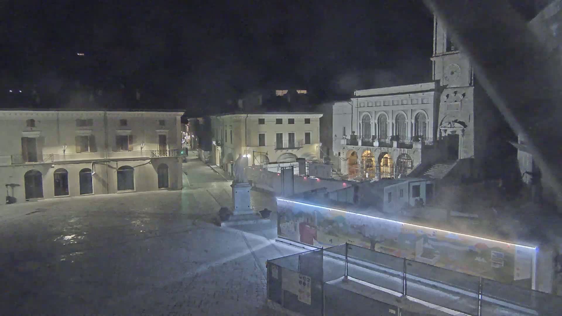 A snow-covered town square at night, featuring several buildings, a statue, and construction barriers with a mural.