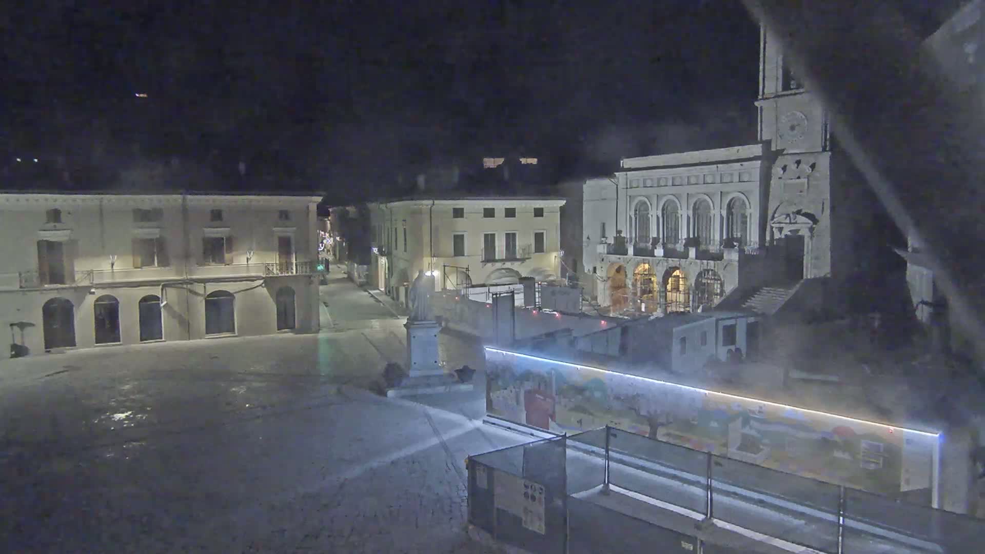 A snow-covered town square at night, with several buildings, a statue, and construction barriers.