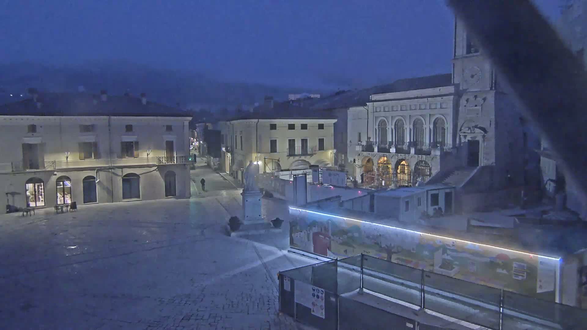 A mostly empty town square at night under a dark, hazy sky, with several buildings surrounding it, one of which is under construction.