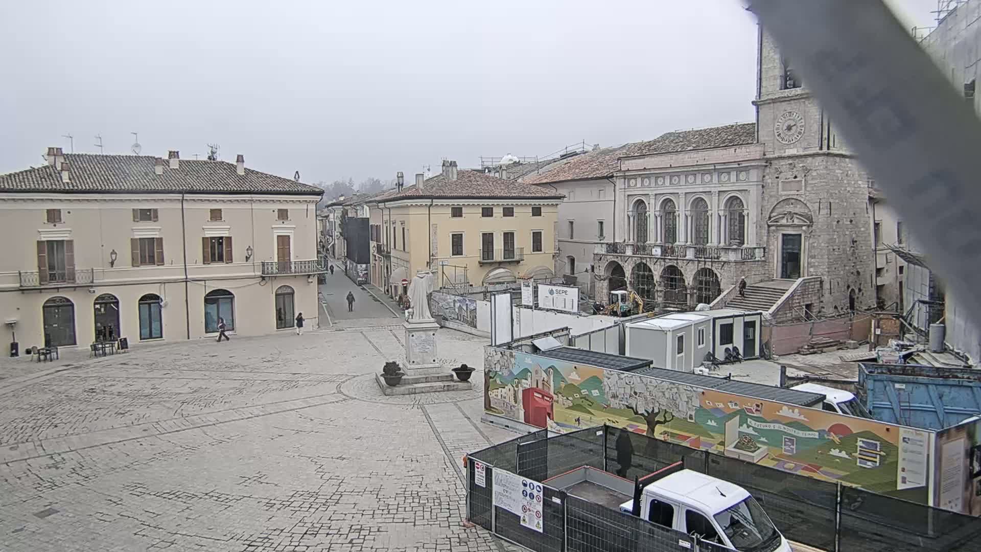 A town square with several buildings, including a large stone building with a clock tower, is under construction, and the weather is overcast.
