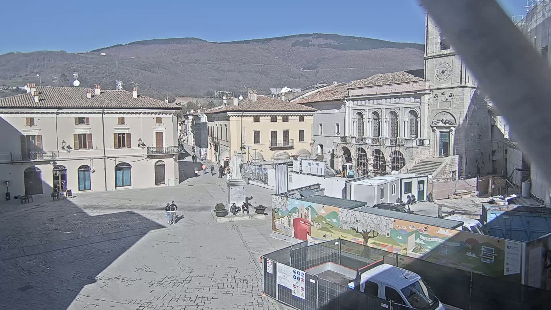 A town square under a clear sunny sky, partially under construction, with several buildings surrounding it and mountains in the background.