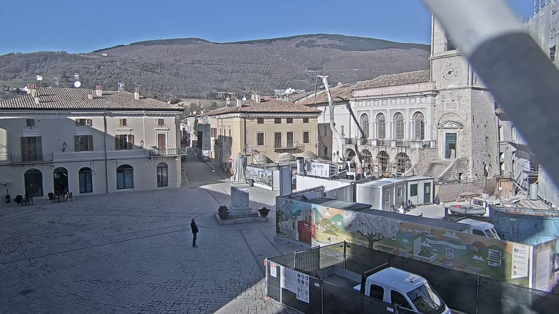 A mostly empty cobblestone town square is under construction, with various construction vehicles and temporary structures present, all in front of older stone buildings and a mountain range under a clear, sunny sky.