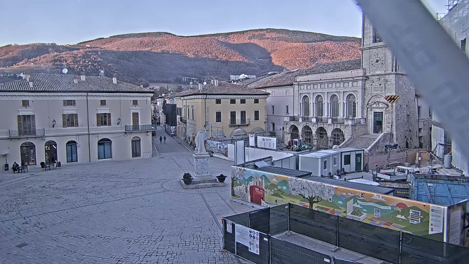 A town square with stone buildings, a statue, and construction underway is visible under a clear, twilight sky with distant mountains in the background.