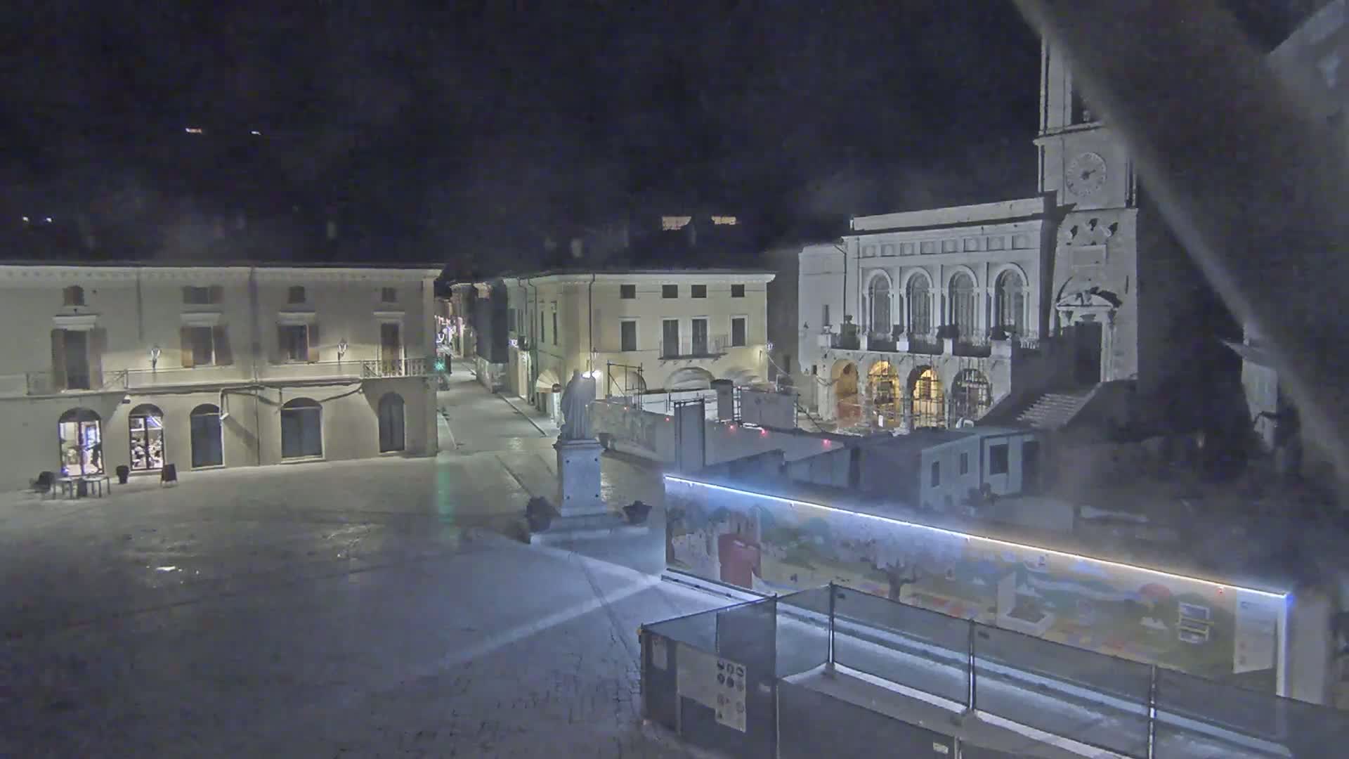 A snow-covered town square at night, featuring several buildings, one of which is a large building with a clock tower, and construction barriers with a mural.