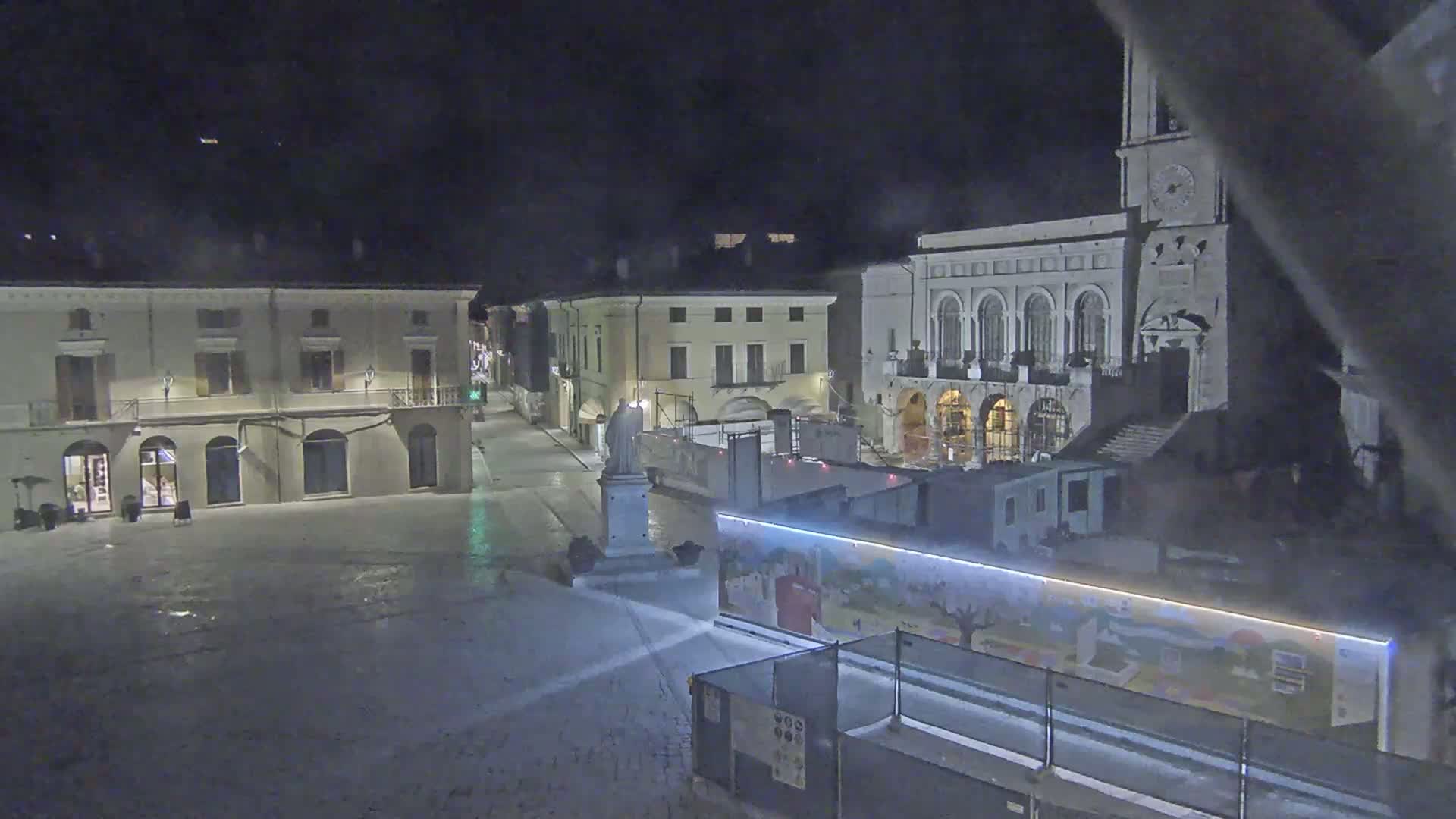 A snow-covered town square at night, featuring several buildings, one of which is a large church with a clock tower, and a construction site with a painted barrier.