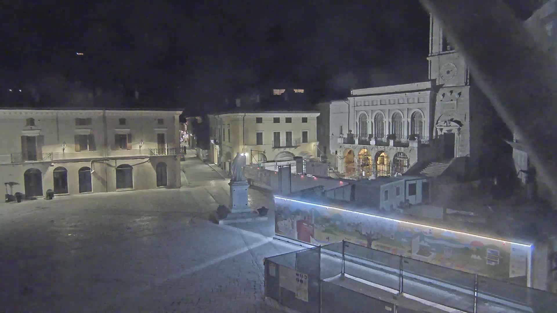 A nighttime view of a town square with several buildings, a statue, and construction barriers illuminated by artificial light.