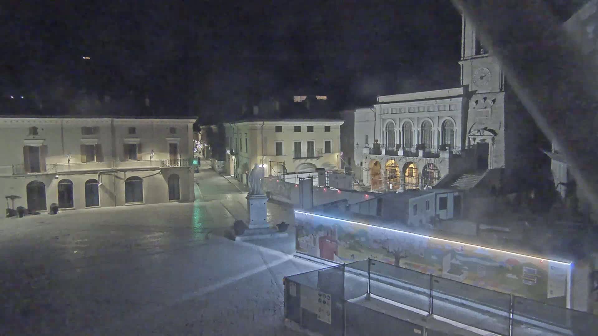 A snow-covered town square at night, featuring several buildings, a statue, and construction barriers with painted artwork.