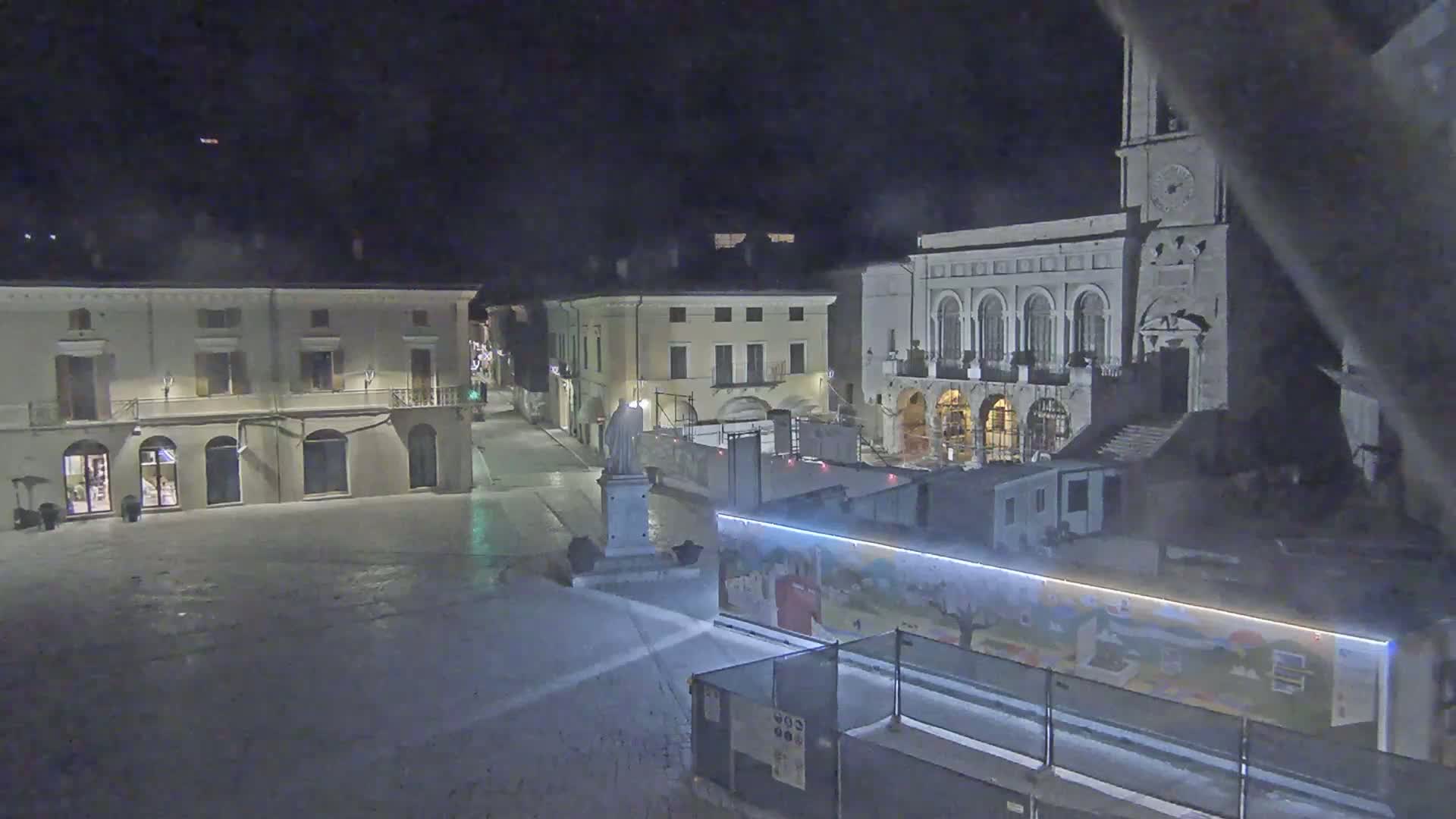 A nighttime view of a town square with several buildings, including one with arched windows and a clock tower, under dark, possibly cloudy conditions.