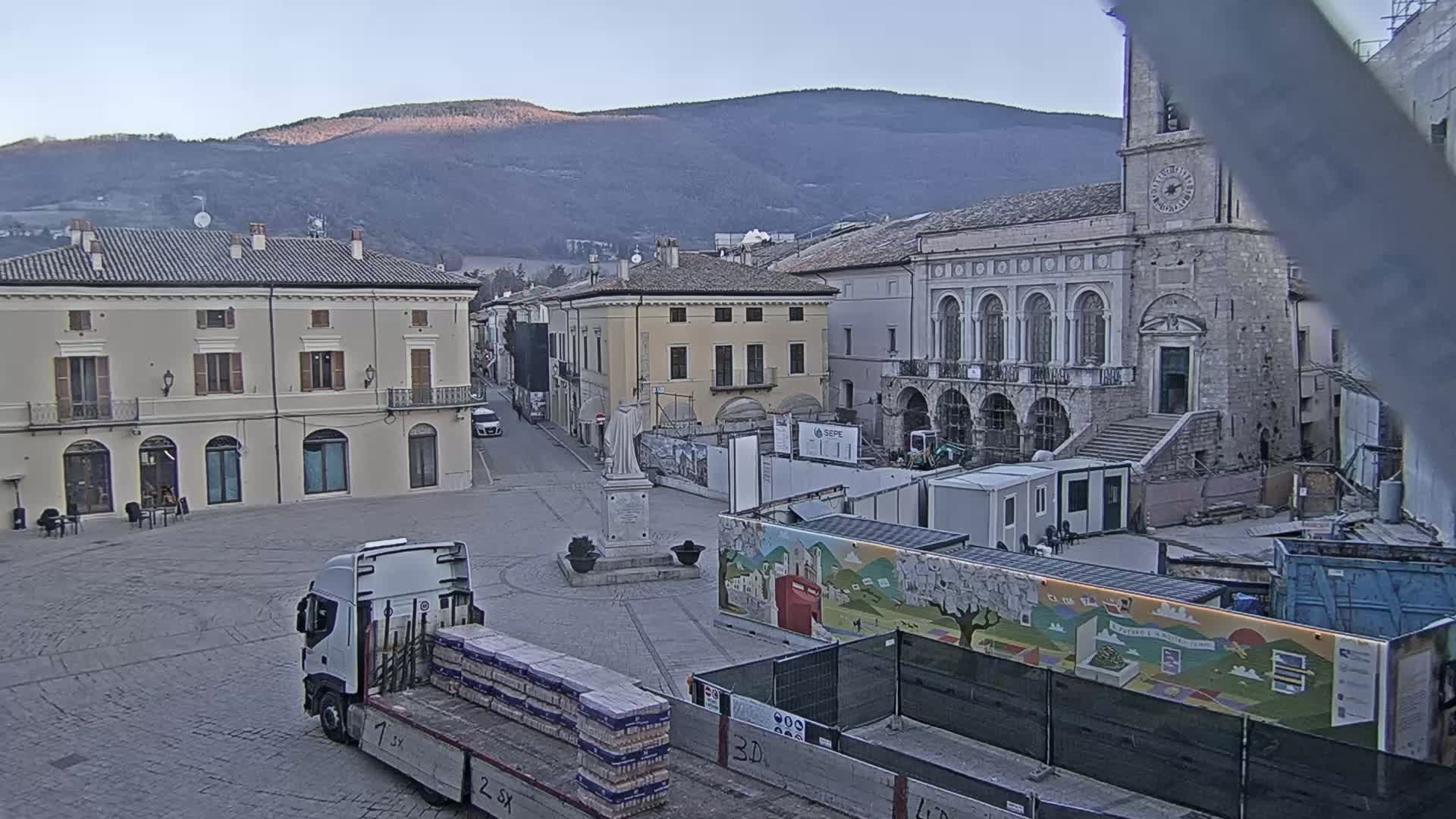 A mostly empty town square with several buildings, including a large stone church, under a clear, light sky with mountains in the background, and construction in progress.