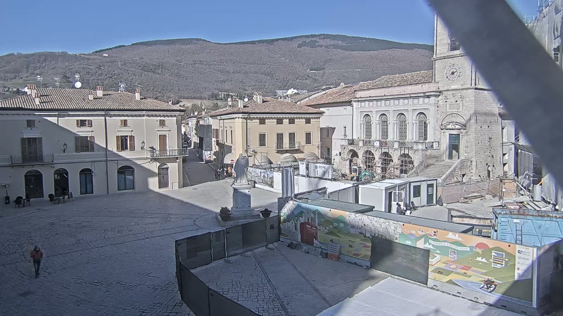 A mostly empty town square is under construction, with buildings surrounding it and mountains in the background on a sunny day.