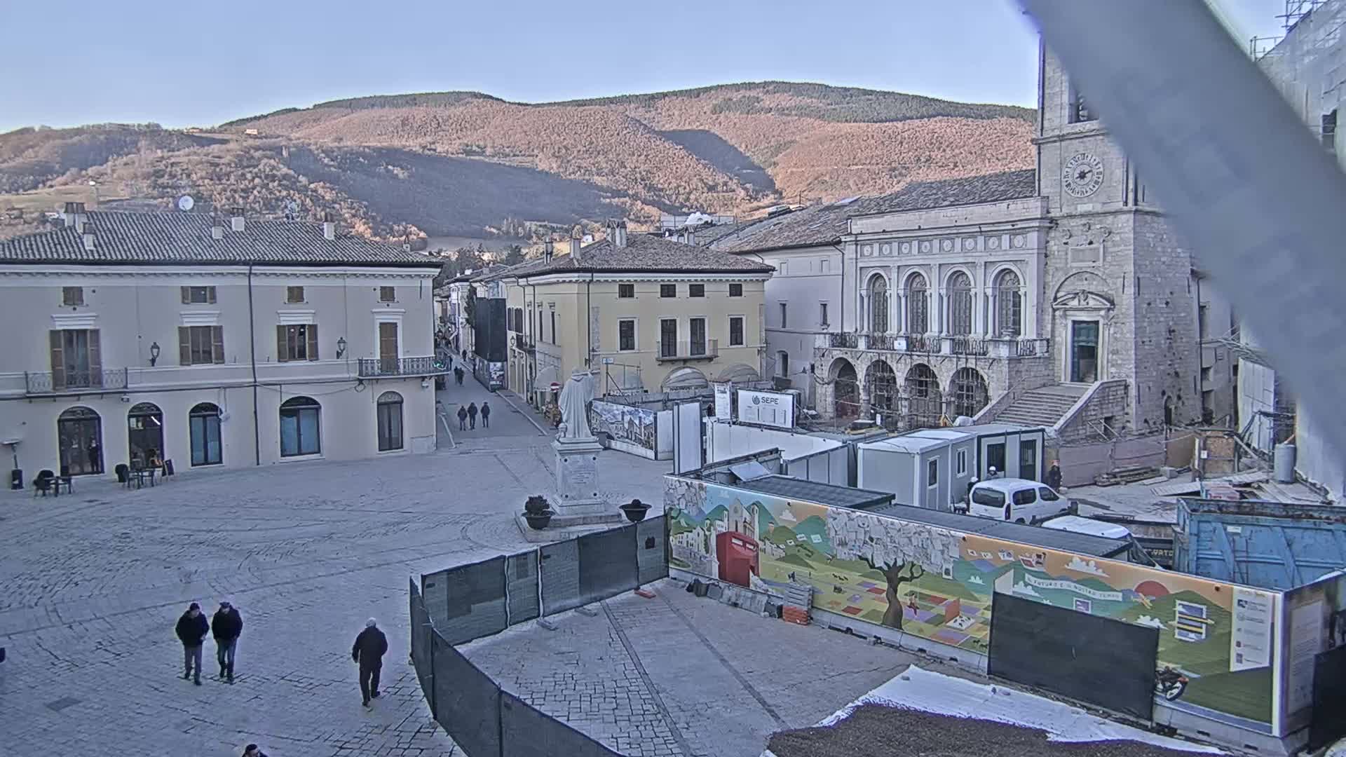 A mostly empty cobblestone town square is under construction, with temporary buildings and fencing surrounding a mural, all situated in front of older buildings and a mountain range under a clear sky.