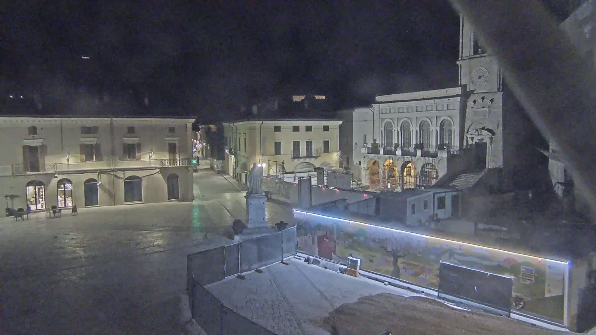 A nighttime view of a town square with several buildings, a statue, and construction barriers, under clear dark skies.