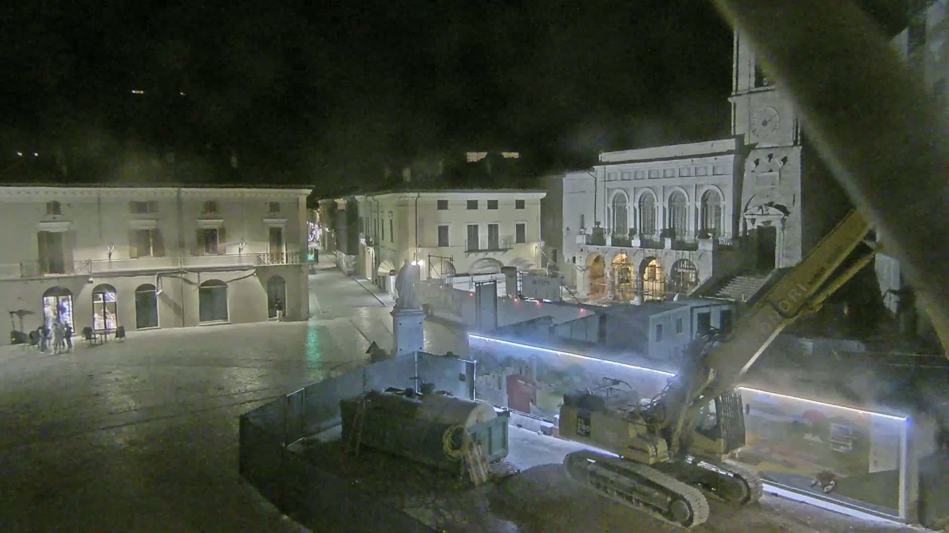 At night, a large excavator works in a town square surrounded by historic buildings under a dark sky.