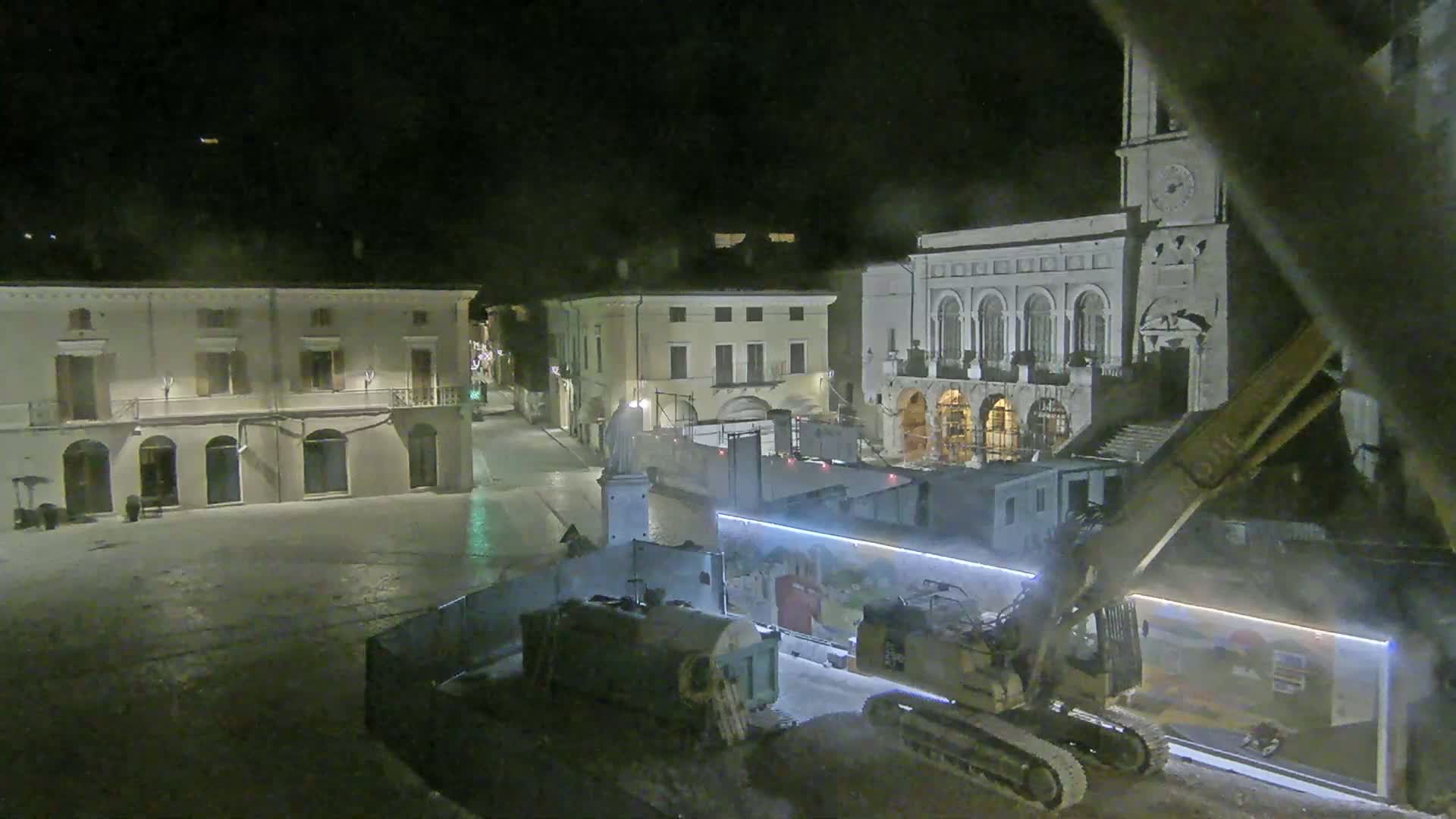 At night, construction equipment operates in a town square surrounded by buildings, under a dark, possibly overcast sky.
