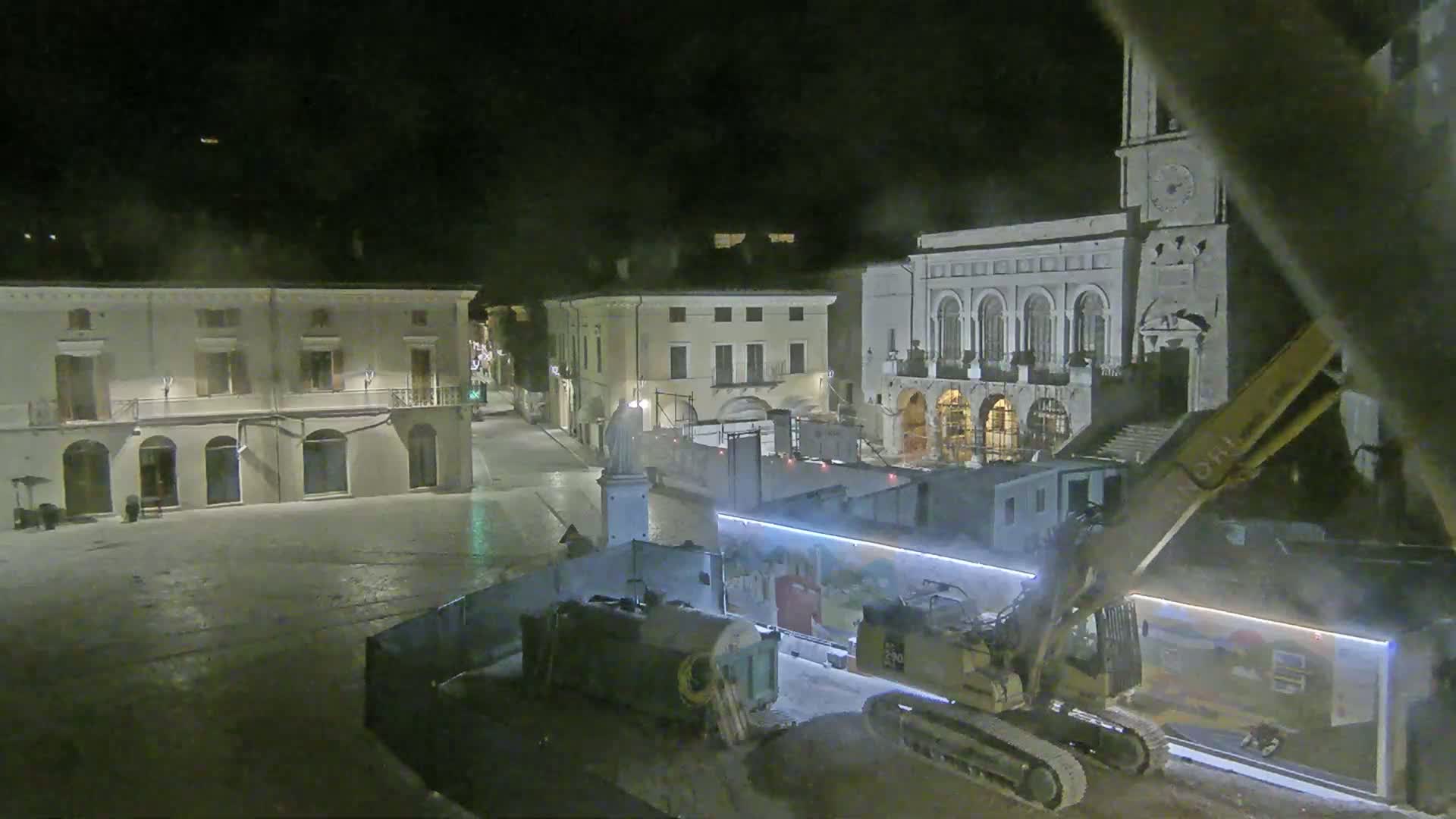 At night, construction equipment is working in a town square surrounded by buildings, under hazy or foggy conditions.