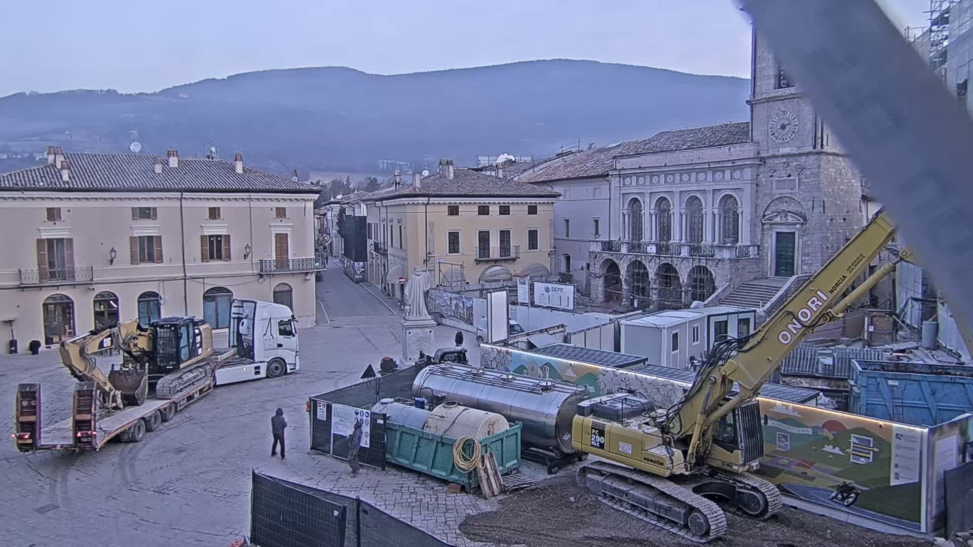 A town square under a hazy sky is undergoing construction, with heavy machinery, trucks, and construction containers present.