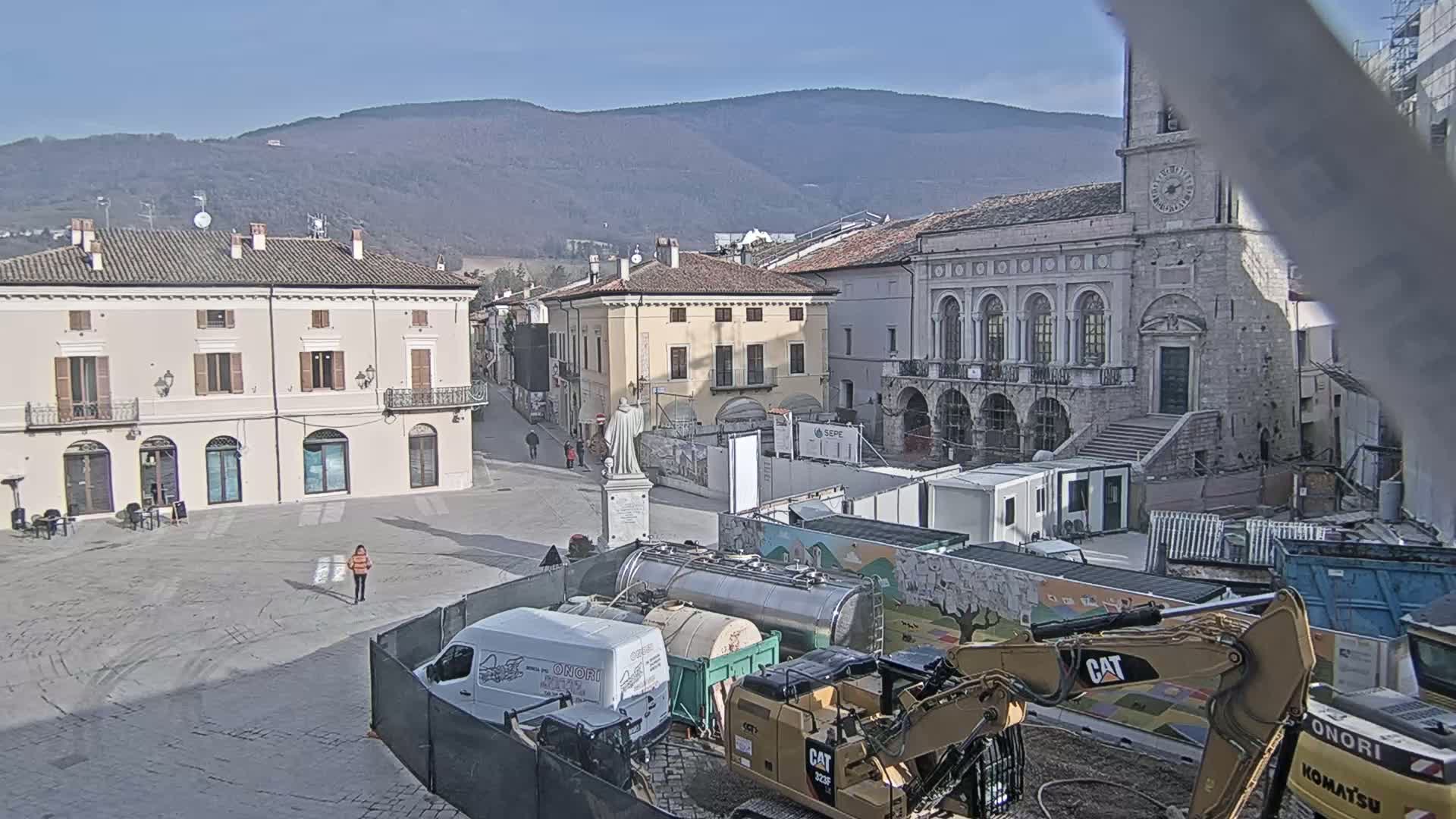 A town square with several buildings, a statue, and construction equipment is visible under sunny skies with mountains in the background.