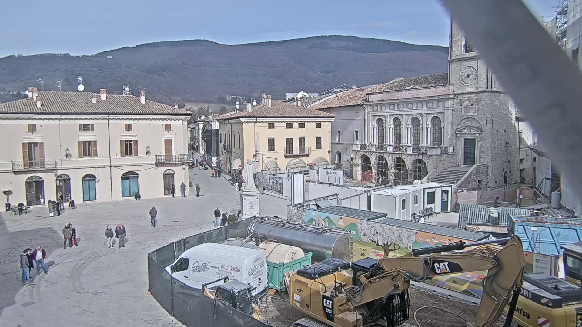 A town square under construction on a sunny day, with construction equipment, temporary buildings, and a few people in the area, backed by historical buildings and mountains in the distance.