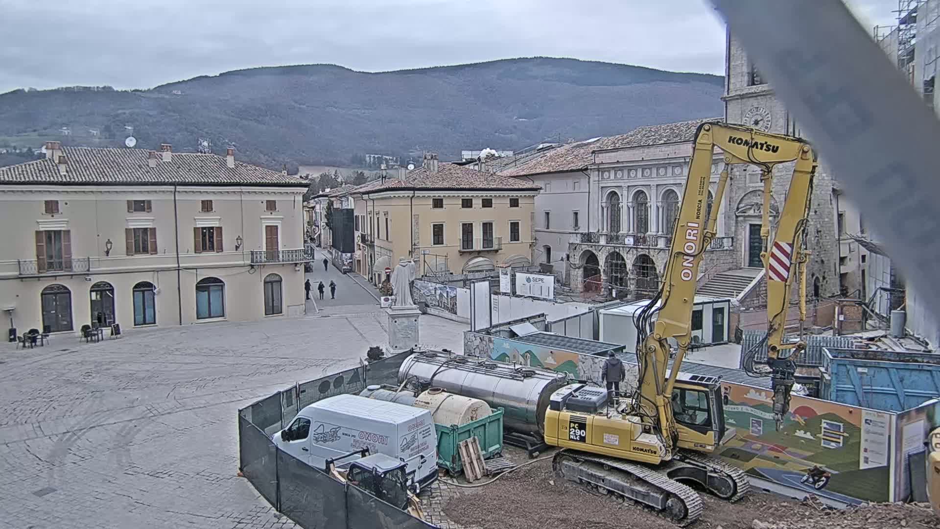 A town square under overcast skies, undergoing construction with an excavator, several vehicles, and temporary fencing present, against a backdrop of buildings and mountains.