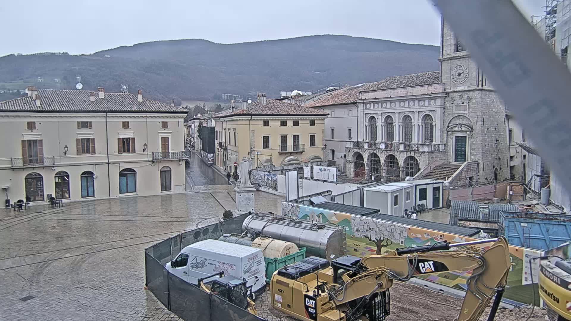 A wet town square under a gray sky, surrounded by old buildings and undergoing construction, with mountains in the background.