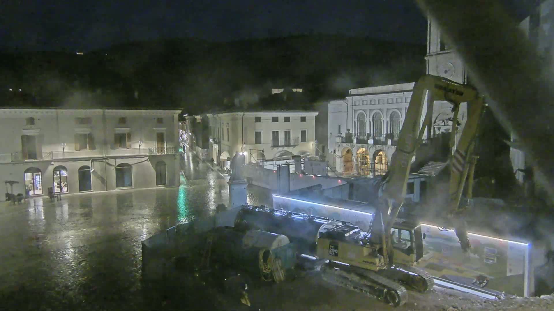 At night, under heavy rain, construction equipment works in a town square near several buildings.