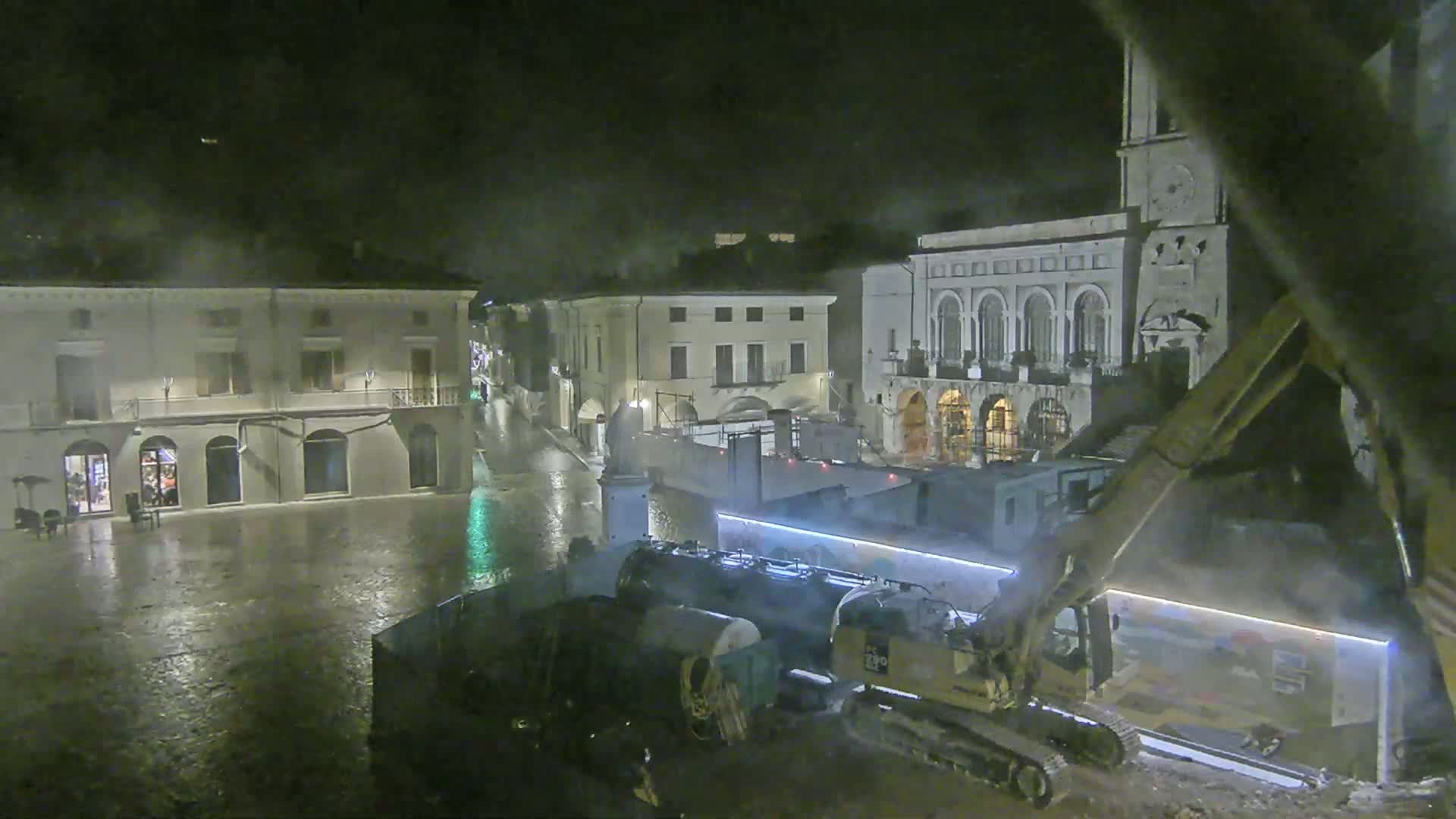 A rain-soaked town square at night is under construction, with several buildings visible and heavy machinery present.