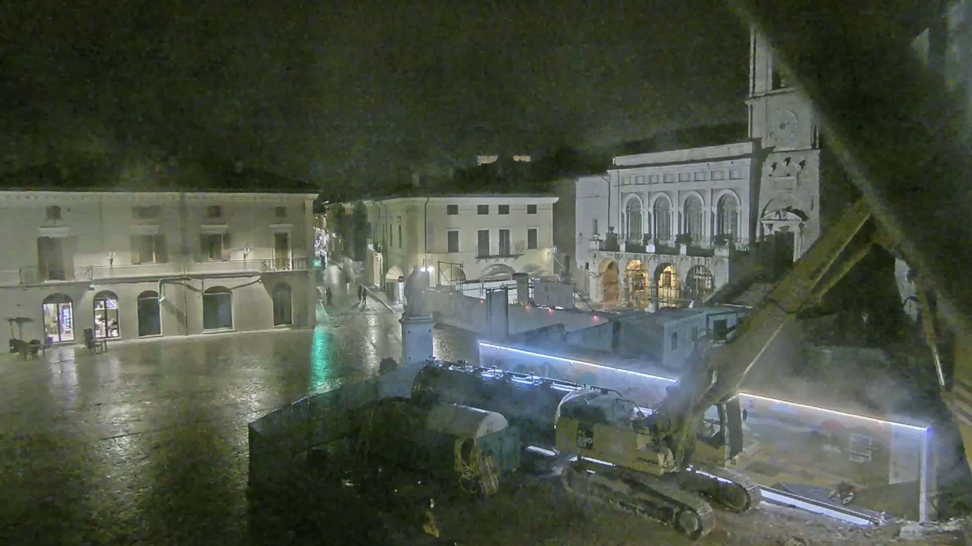 A nighttime view of a town square, under rainy conditions, shows construction equipment near several buildings, one with a clock tower.