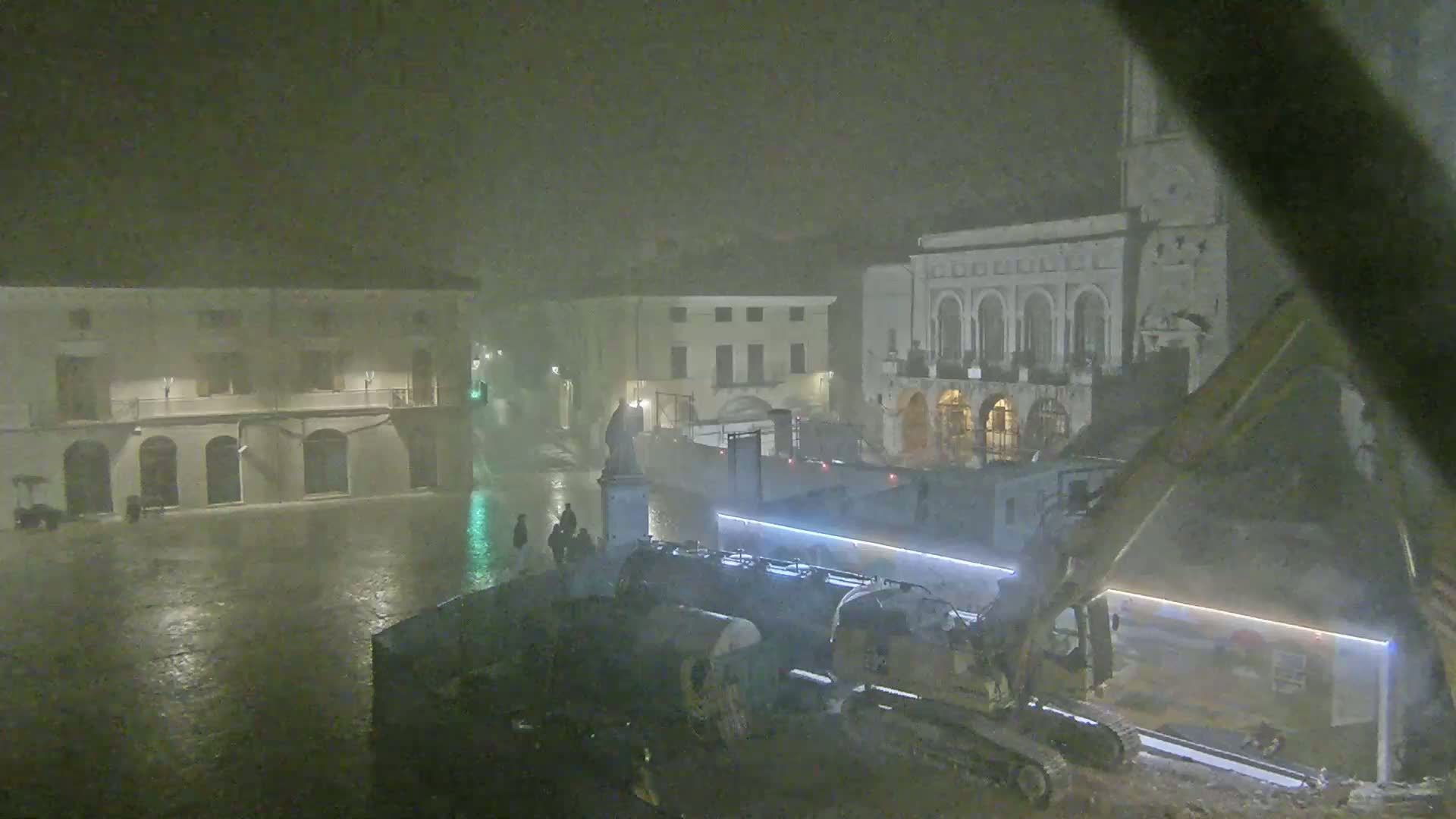 A rain-swept town square at night shows construction equipment near buildings, with a statue visible in the background.