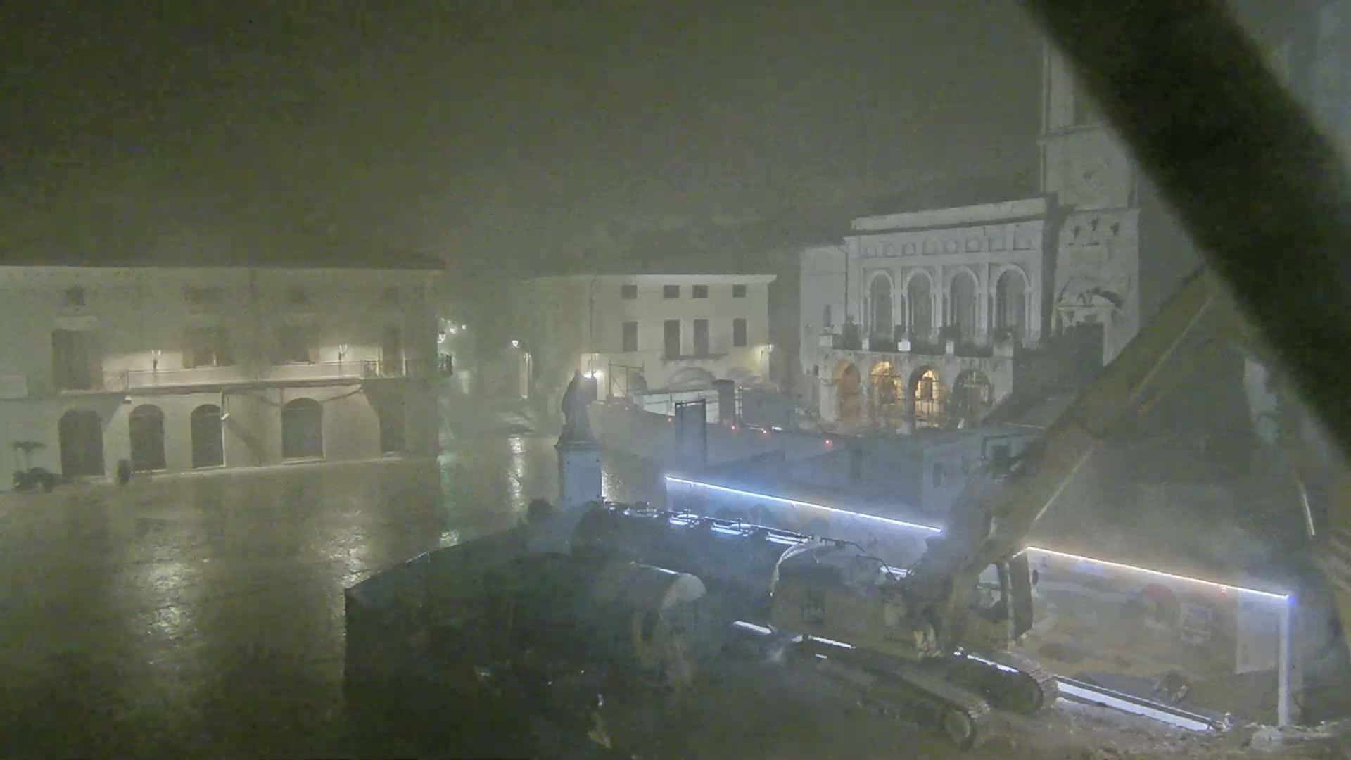 A rain-soaked town square at night is shown with construction equipment and several buildings, one of which is a large, ornate structure.